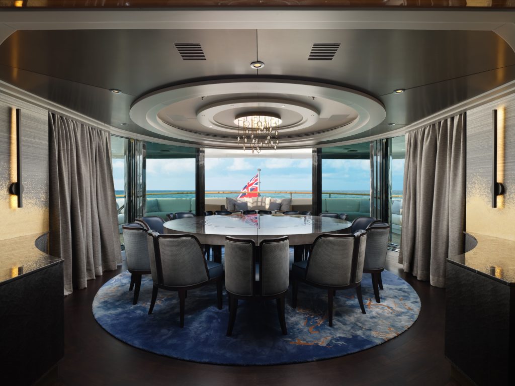 A luxurious dining room with a large round table, upholstered chairs, modern lighting, and floor-to-ceiling windows overlooking the ocean. A British flag is visible outside on the balcony.