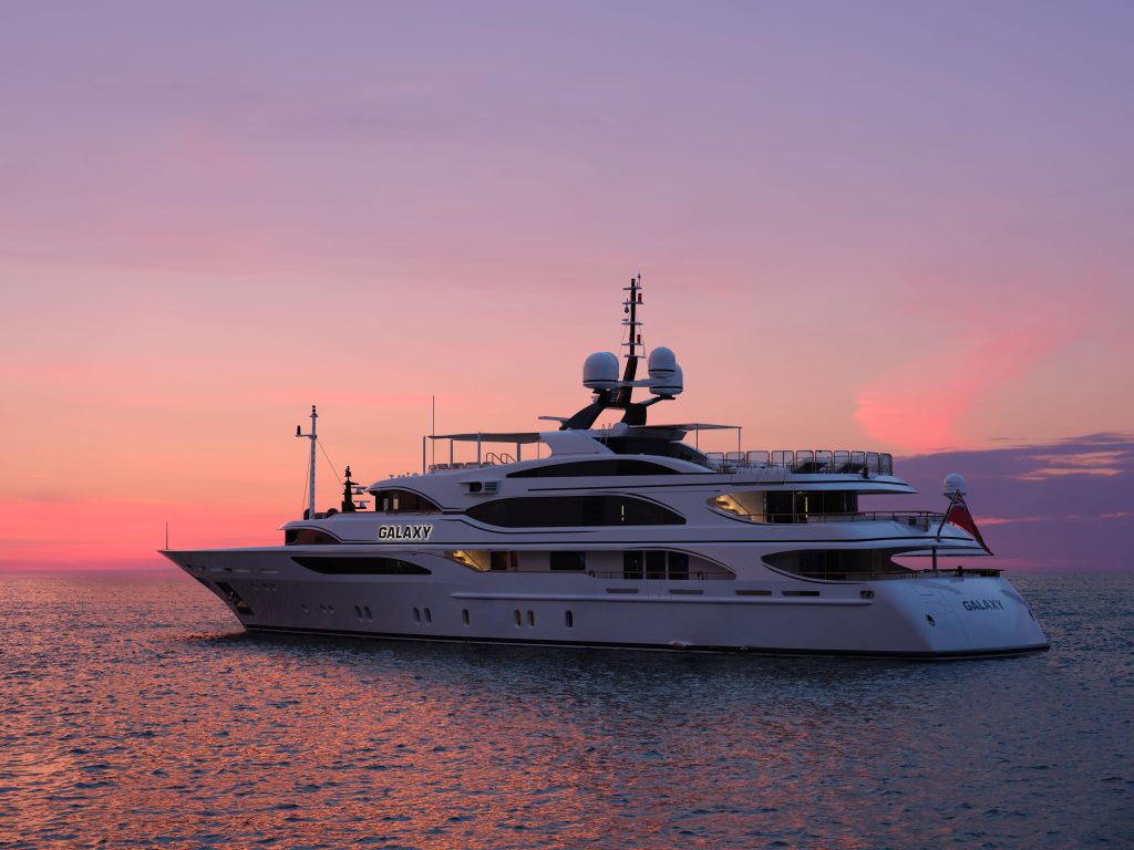 A luxury yacht named GALAXY floats on calm water at sunset, with a vibrant pink and purple sky in the background.