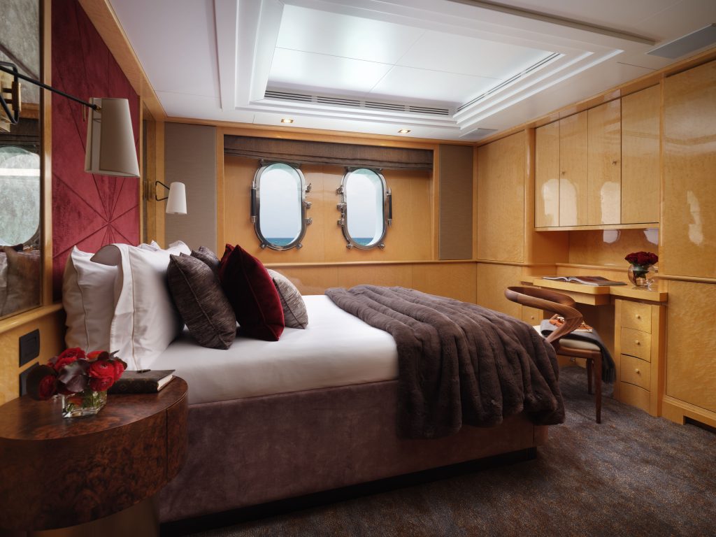 A cozy, modern cruise ship cabin features a double bed with plush pillows and a dark throw, two round porthole windows, a wooden desk with a chair, built-in cabinets, and soft lighting. Flowers decorate the bedside table and desk.