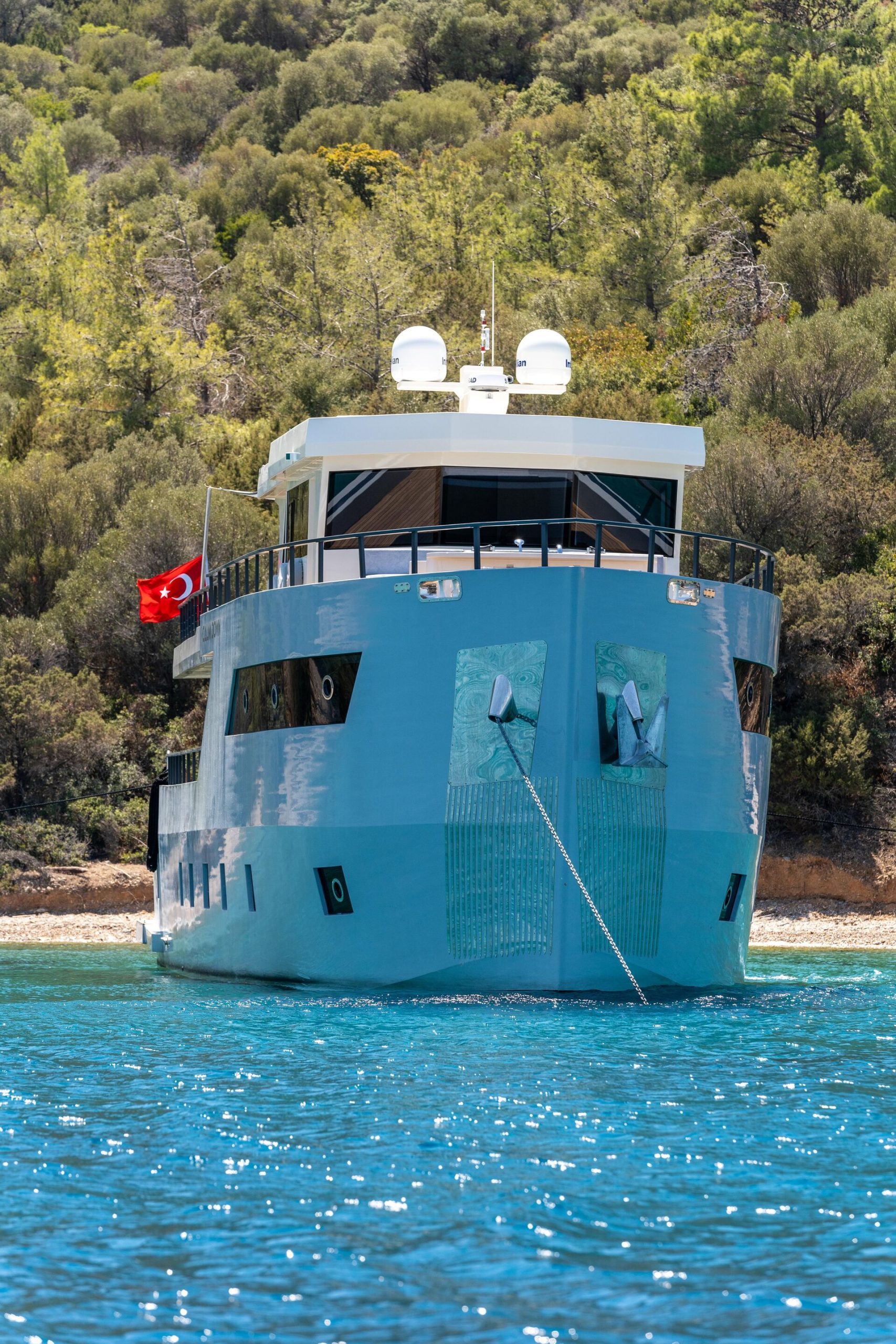 A modern yacht with a Turkish flag anchored near a forested shore, reflecting blue water under a clear sky.