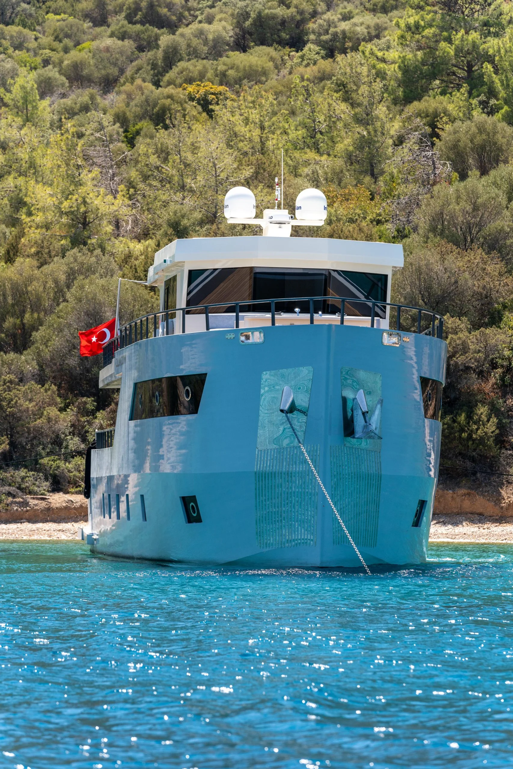 A modern yacht with a Turkish flag anchored near a forested shore, reflecting blue water under a clear sky.