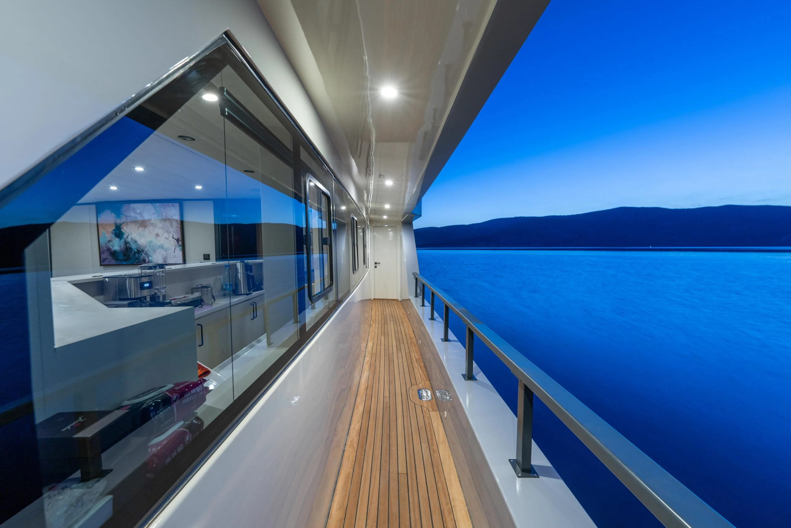A modern yacht’s exterior walkway with wooden flooring, overlooking calm blue water and distant mountains at dusk. Interior lights reflect off large windows showing a kitchen inside.