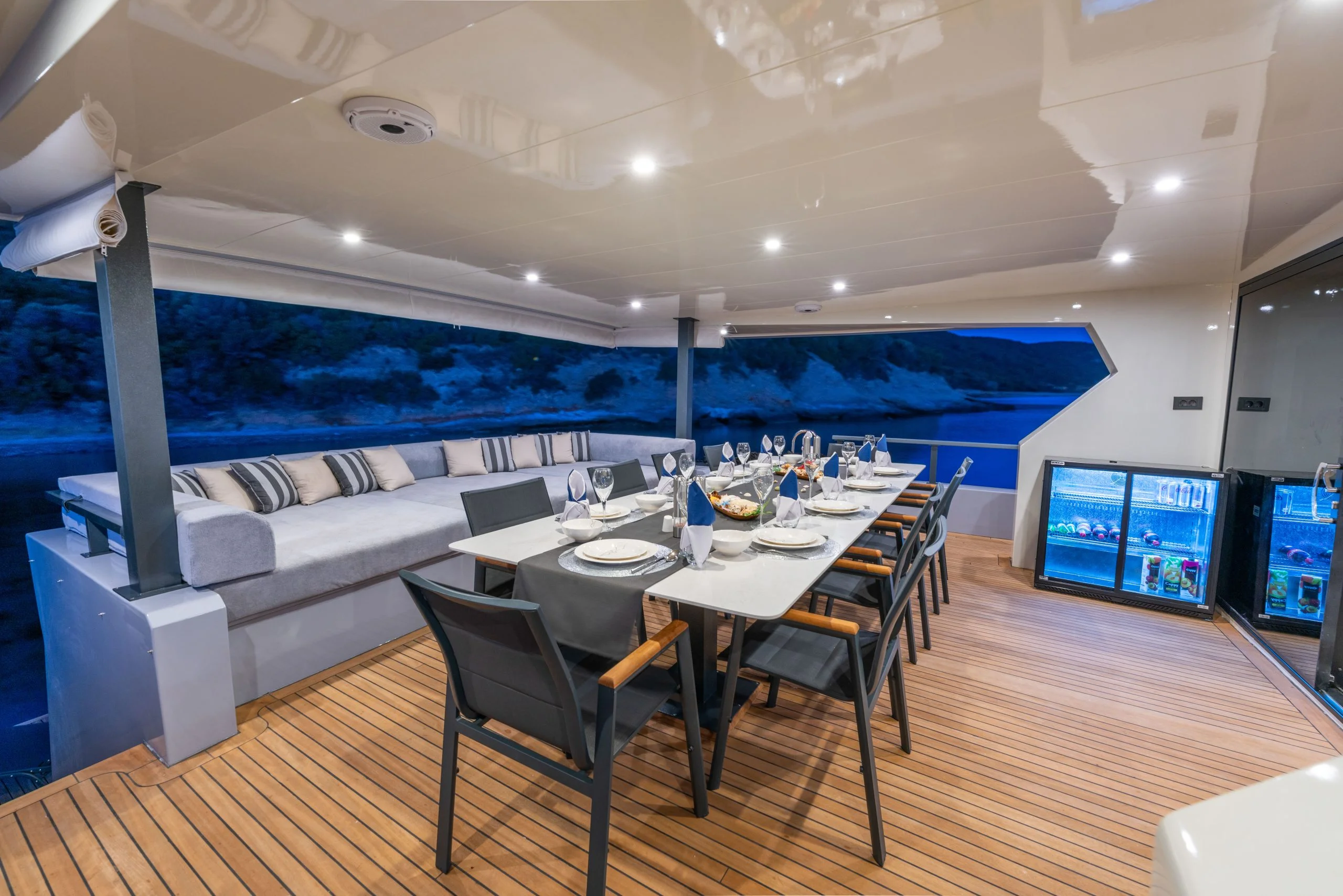 Luxurious yacht deck with a large cushioned seating area, a dining table set for eight, and a mini fridge, overlooking water and hills in the evening. Warm recessed lighting creates a cozy atmosphere.