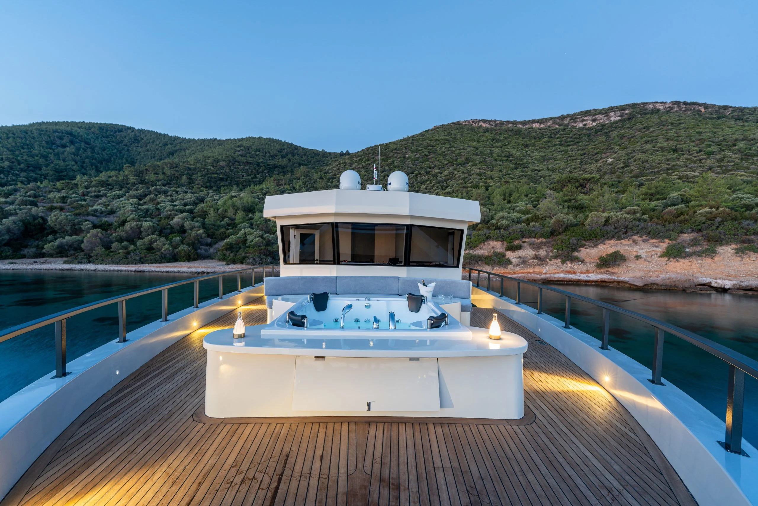 A luxury yacht deck with a jacuzzi in the center, surrounded by glass railings, overlooking calm water and tree-covered hills at sunset.