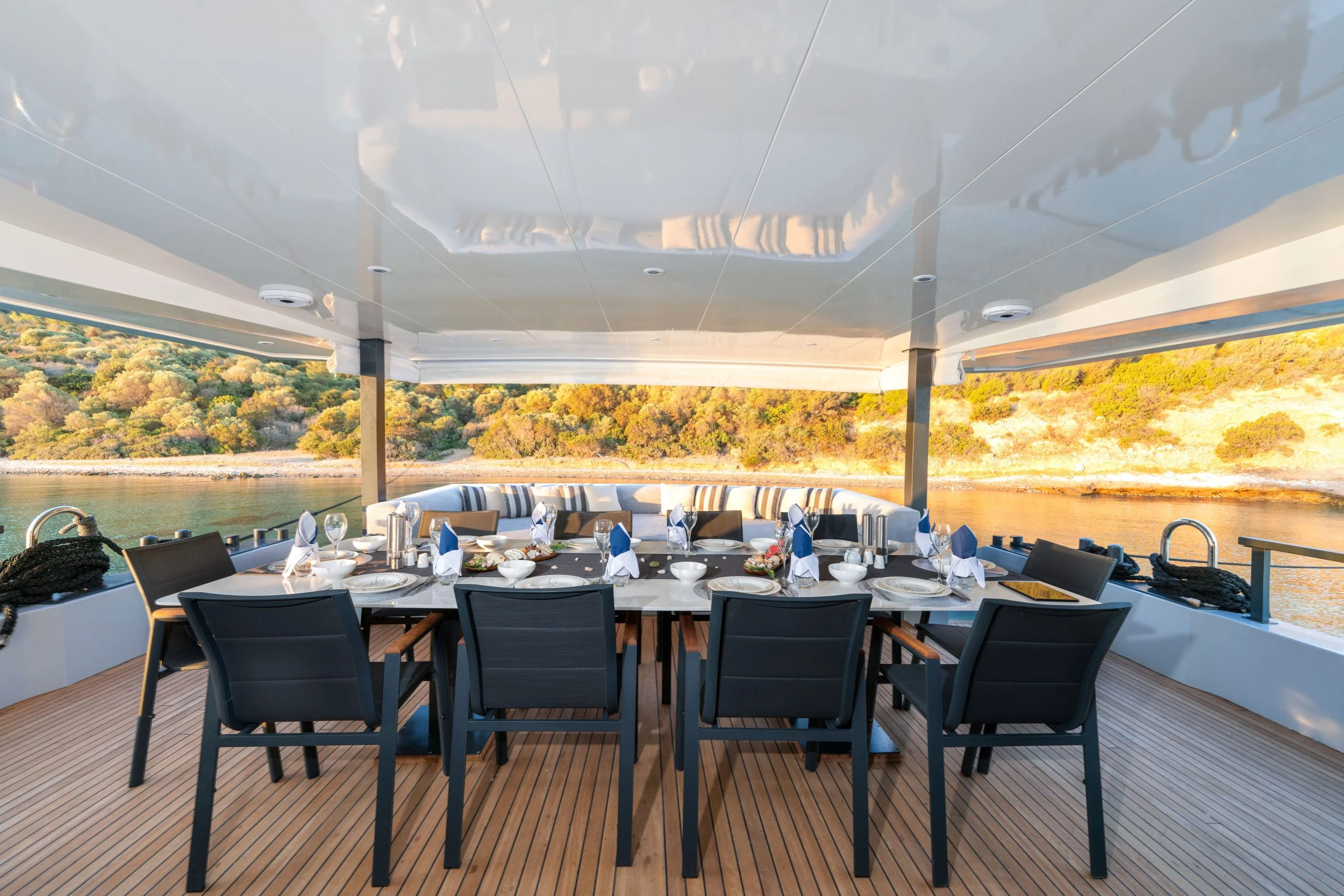 Elegant outdoor dining table set for ten on a yacht deck, overlooking calm water and a sunlit, tree-lined shore in the background. The table is arranged with plates, glasses, and napkins.