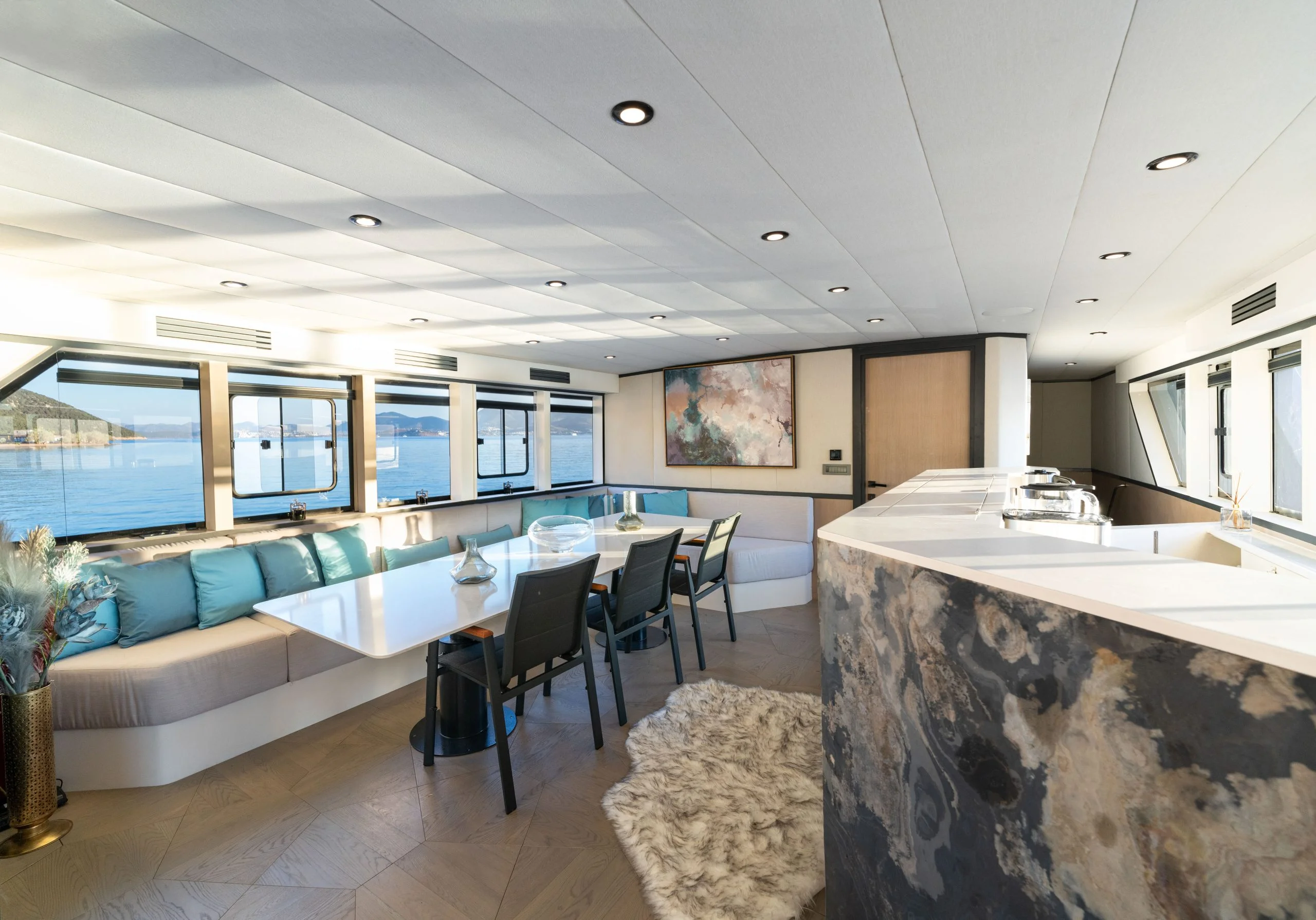 A modern yacht interior with large windows, a dining table with chairs, a cushioned seating area, coastal decor, and a kitchen counter. Sunlight fills the space, highlighting the view of the water outside.