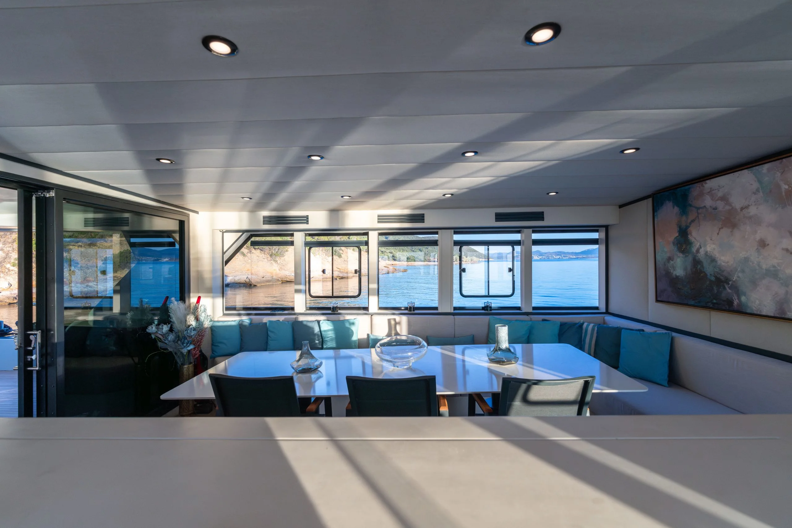 A modern yacht interior with a large white dining table, turquoise cushions on a built-in bench, glass doors, large windows with a sea view, and a painting on the wall. Sunlight streams into the room.