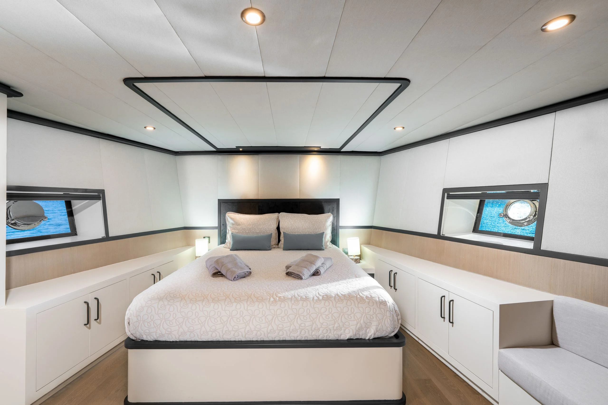 Modern yacht bedroom with a double bed, two rolled towels on top, bedside lamps, porthole windows on each side, and built-in white cabinetry along the walls. The room has clean lines and bright lighting.