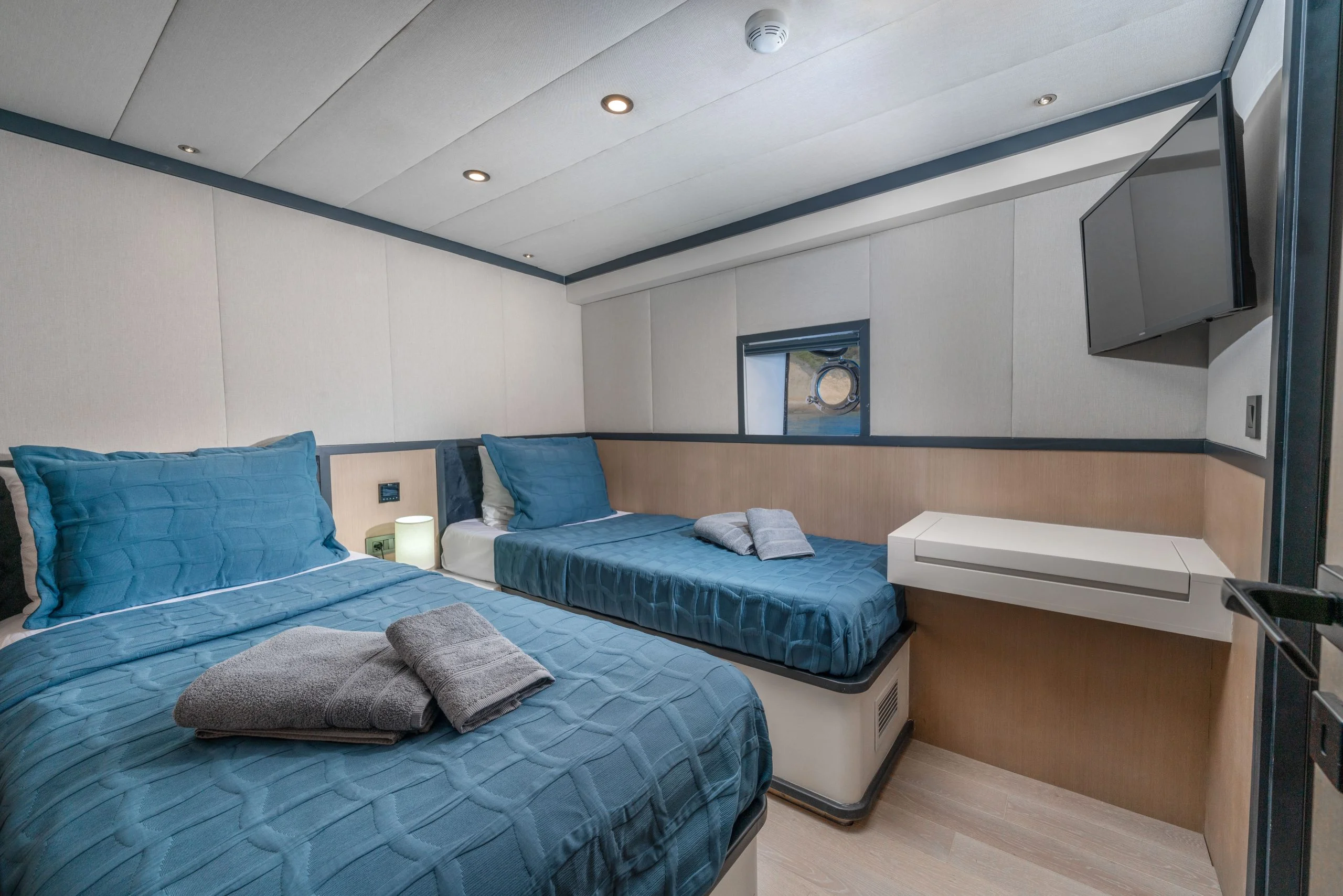 A modern yacht cabin with two single beds made up in blue bedding, a small round window, a wall-mounted TV, a floating desk, and neatly folded towels placed on each bed.