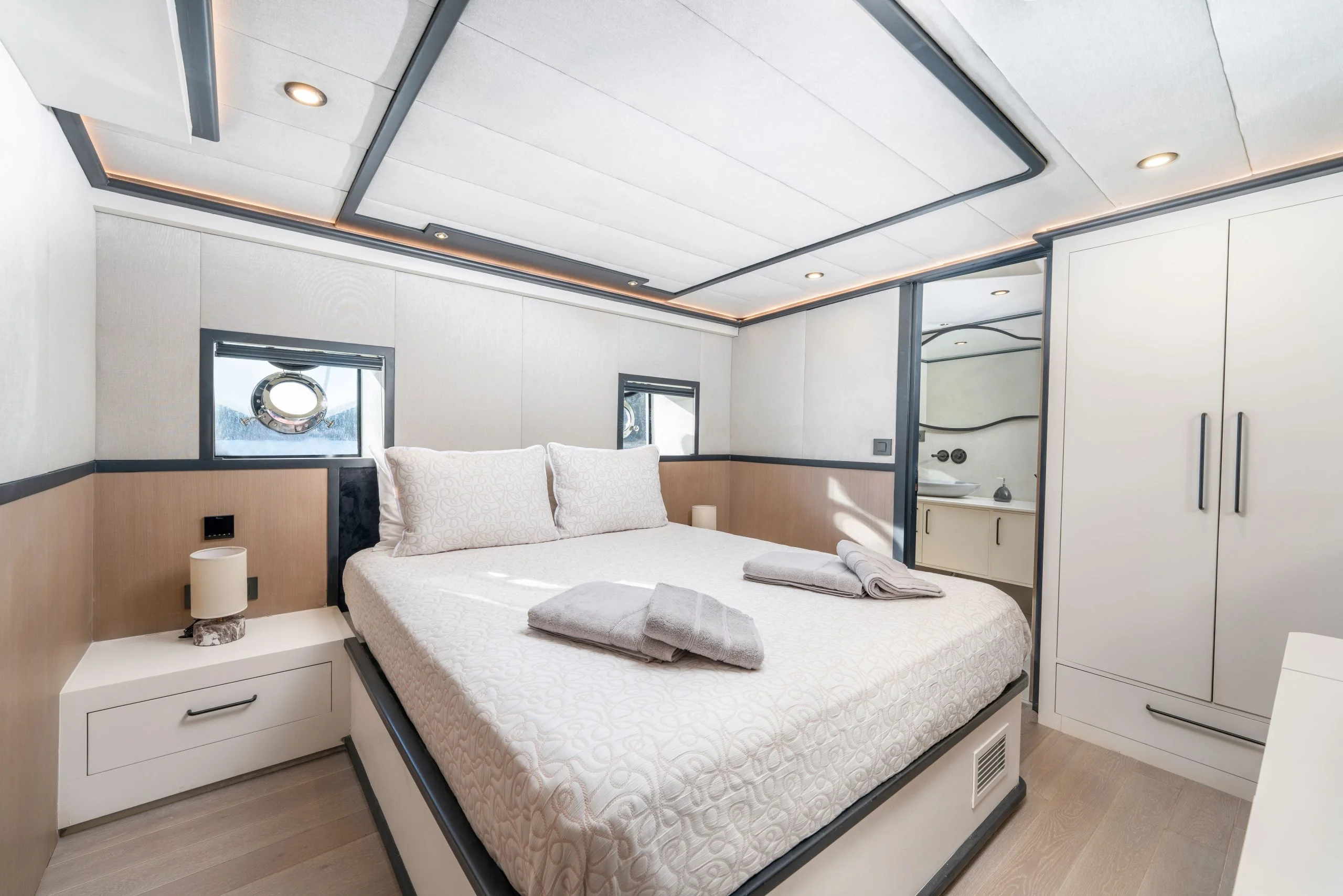 Modern yacht bedroom with a large bed, light wood and white decor, two bedside tables, towels on the bed, small round windows on the walls, and a view into a bathroom with a sink and mirror.