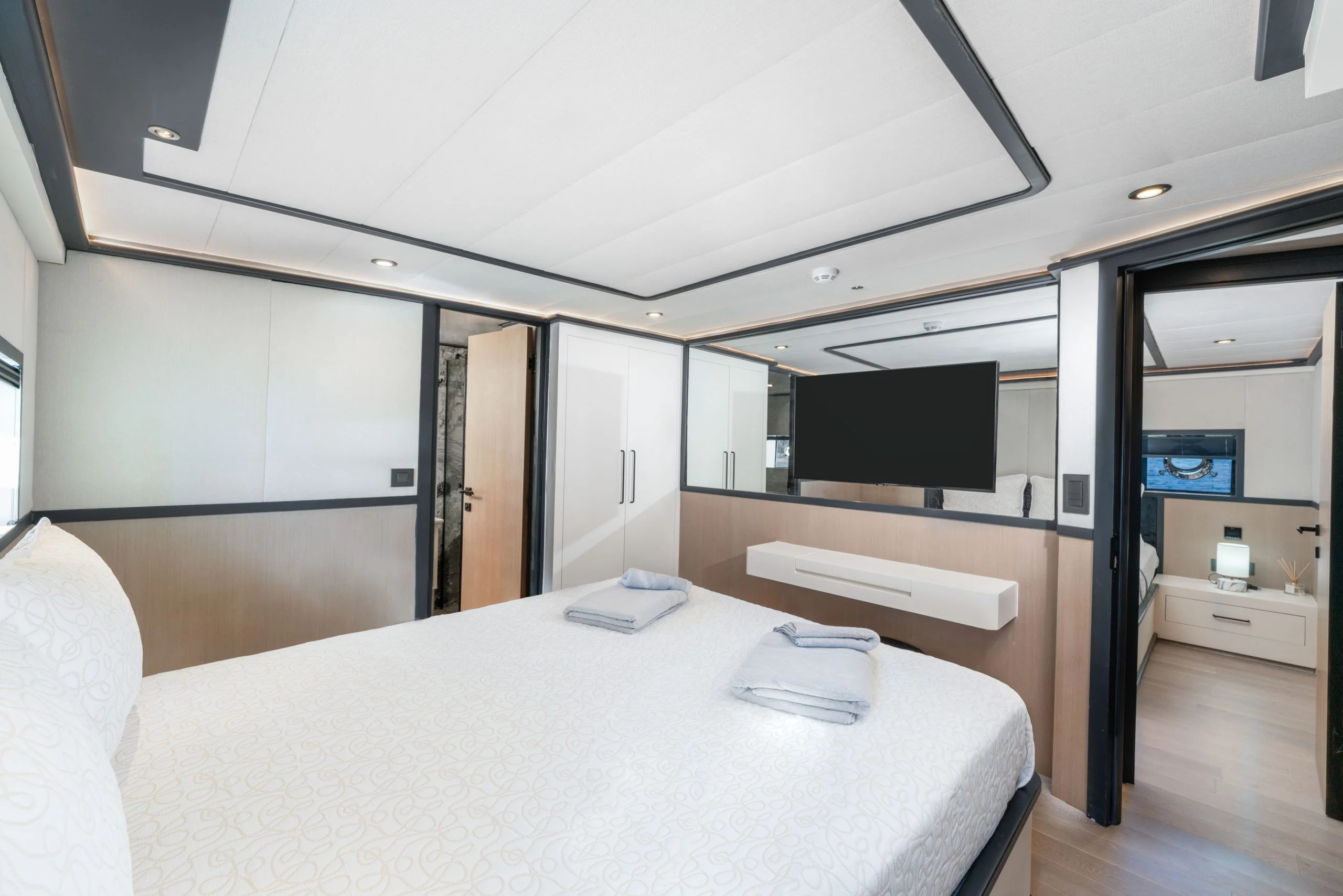 Modern bedroom with a large bed, white bedding, neatly folded towels, a wall-mounted TV, built-in wardrobe, and a view into an adjoining room with a nightstand and lamp.