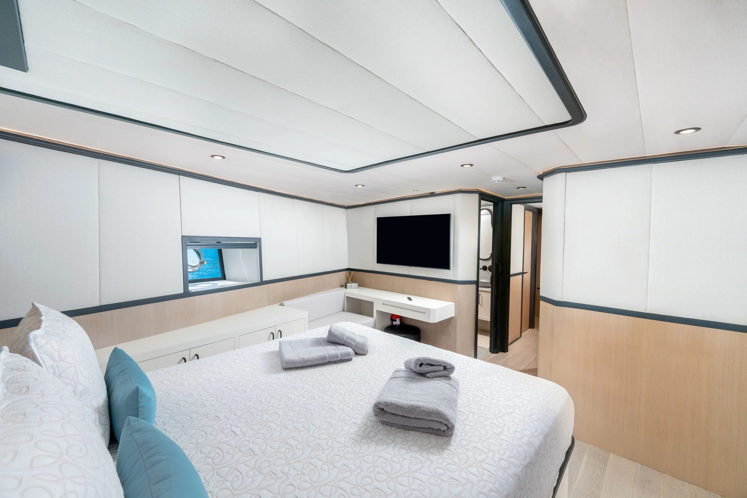 Modern yacht bedroom with a large bed, decorative pillows, and neatly folded towels. The room has white walls, built-in storage, a wall-mounted TV, a small window, and an ensuite bathroom in the background.