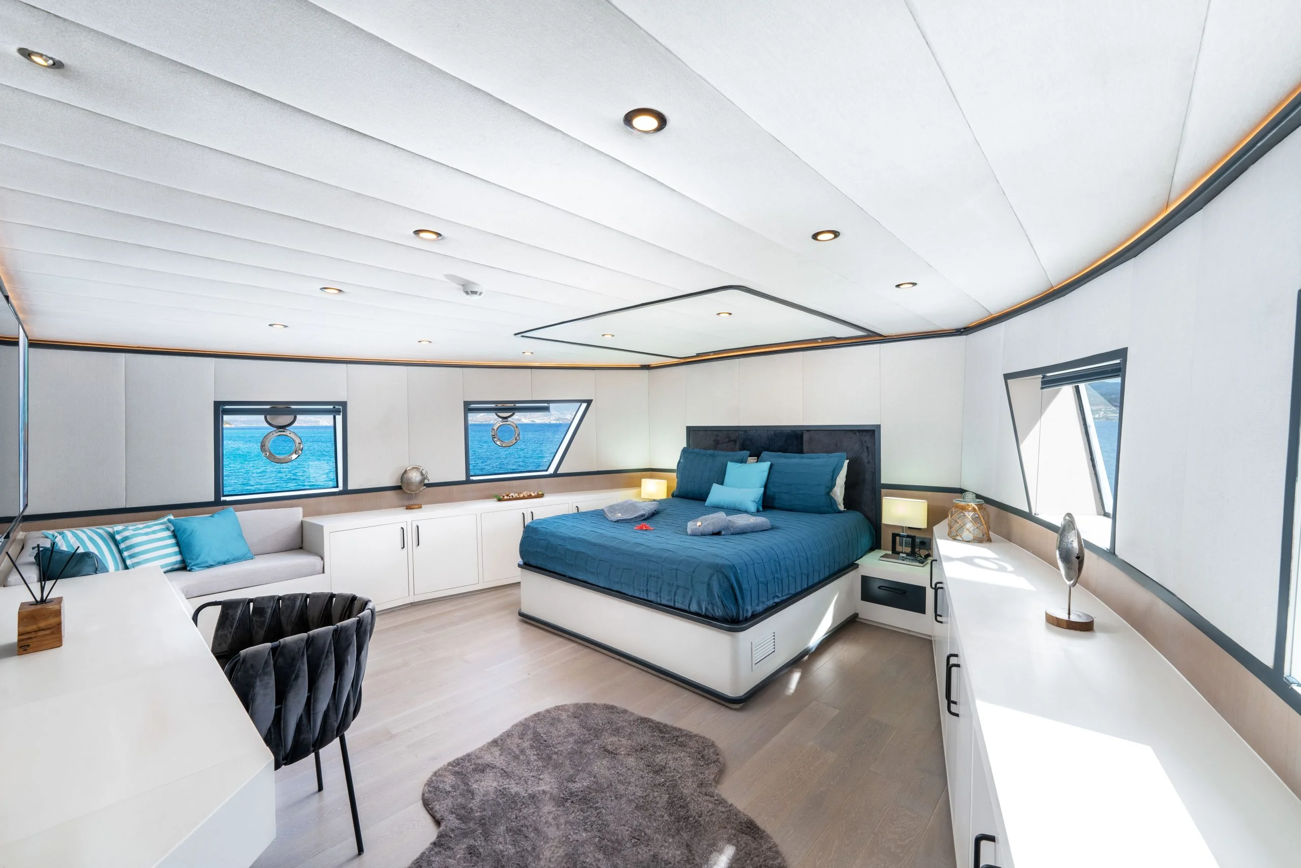 A modern yacht bedroom with large windows, a blue bed, white furniture, a small couch with striped pillows, built-in storage, and ocean views. The room is bright, spacious, and decorated in blue and white tones.