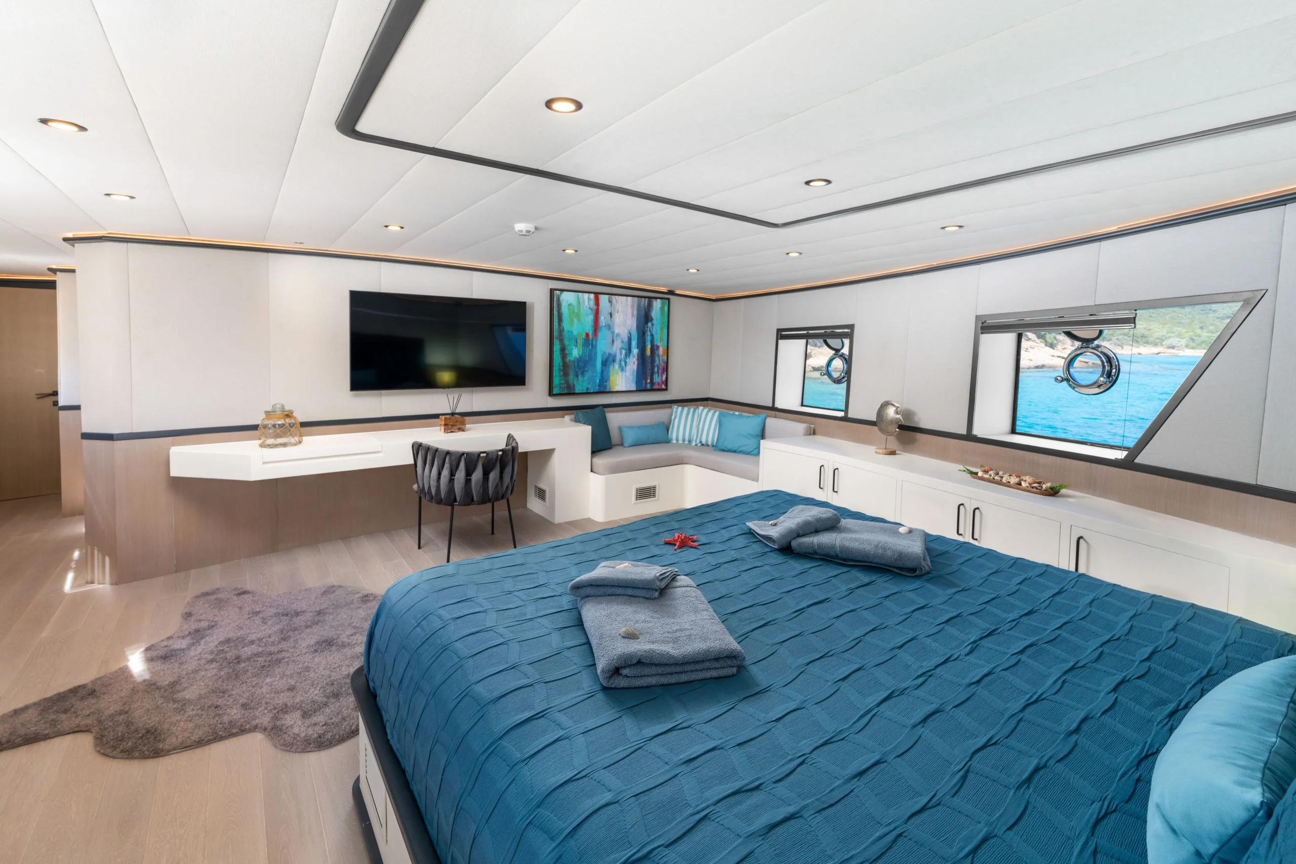 A spacious yacht bedroom with a large bed covered in a blue blanket, two folded towels, a desk, TV, and windows overlooking the ocean. The room is modern and bright with nautical decor.