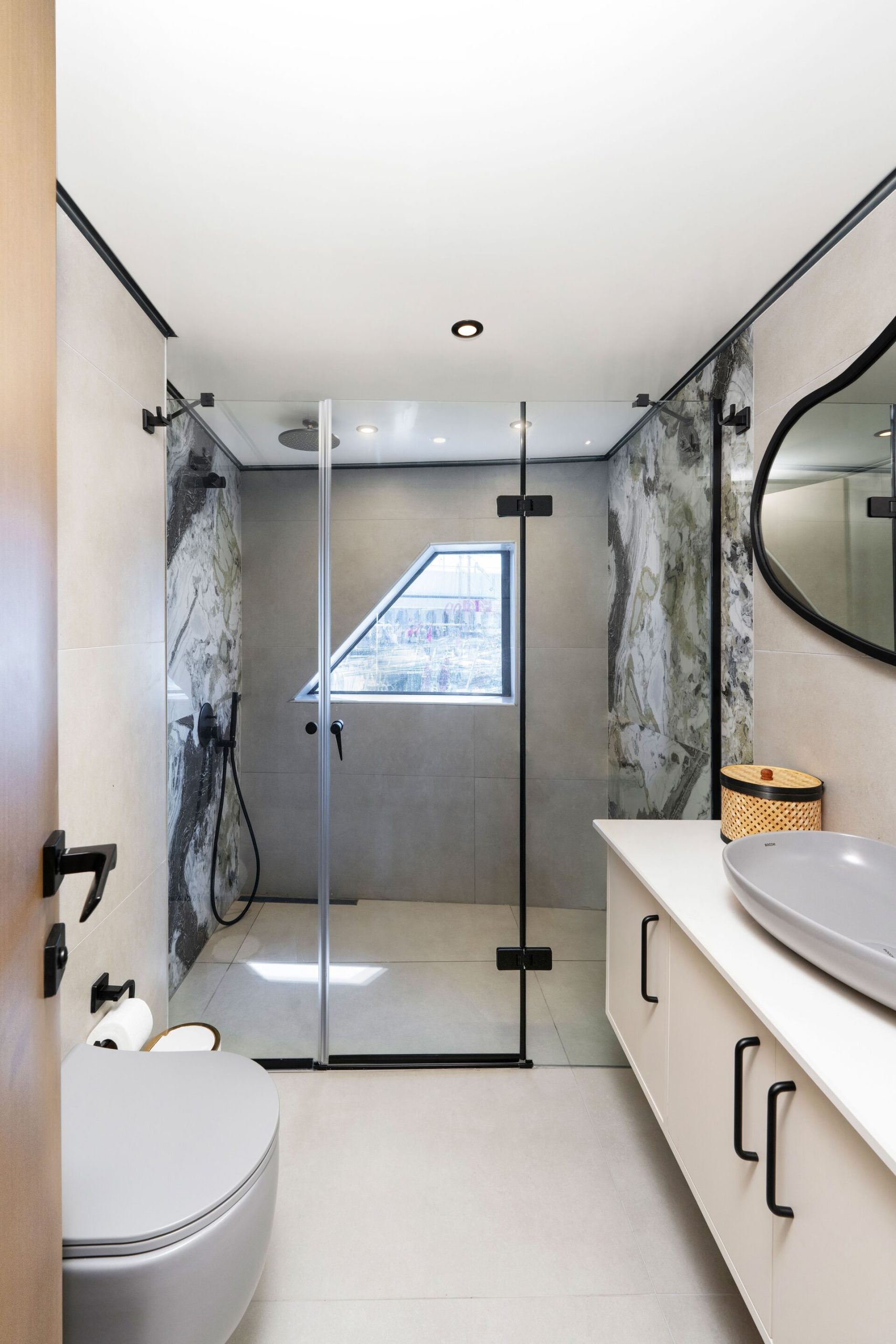 Modern bathroom with a glass-enclosed shower, wall-mounted toilet, large oval mirror, vessel sink on a white countertop, and a window letting in natural light above a grey and white marble wall.