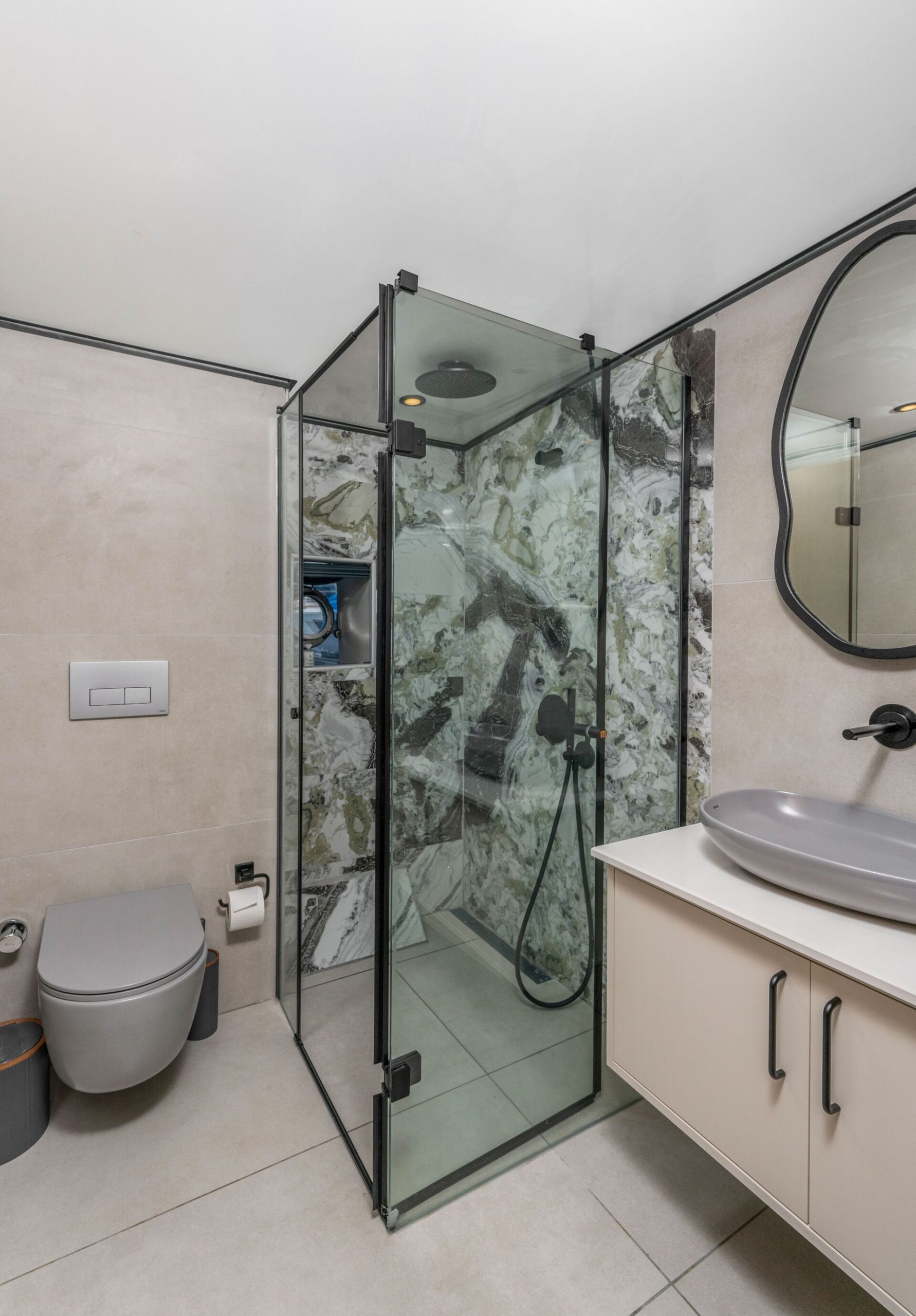 Modern bathroom with a glass shower enclosure featuring a marbled wall, wall-mounted toilet, oval mirror, floating vanity with a gray vessel sink, and minimalist fixtures.