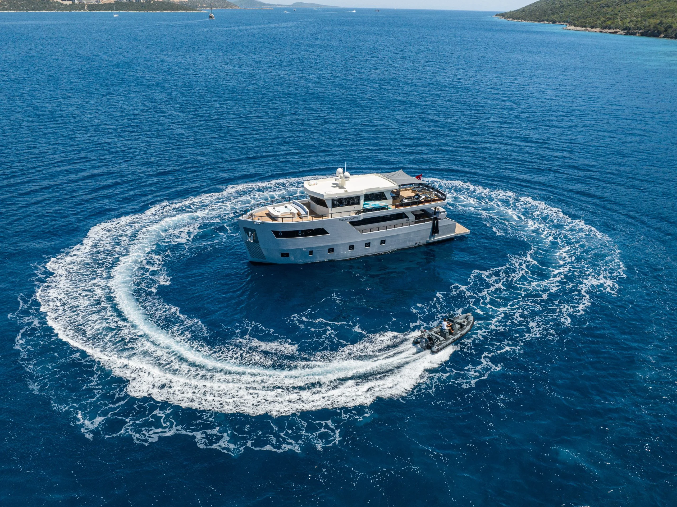 A motor yacht is anchored in clear blue water while a small speedboat creates a circular wake around it. Lush green coastline is visible in the distance under a clear sky.