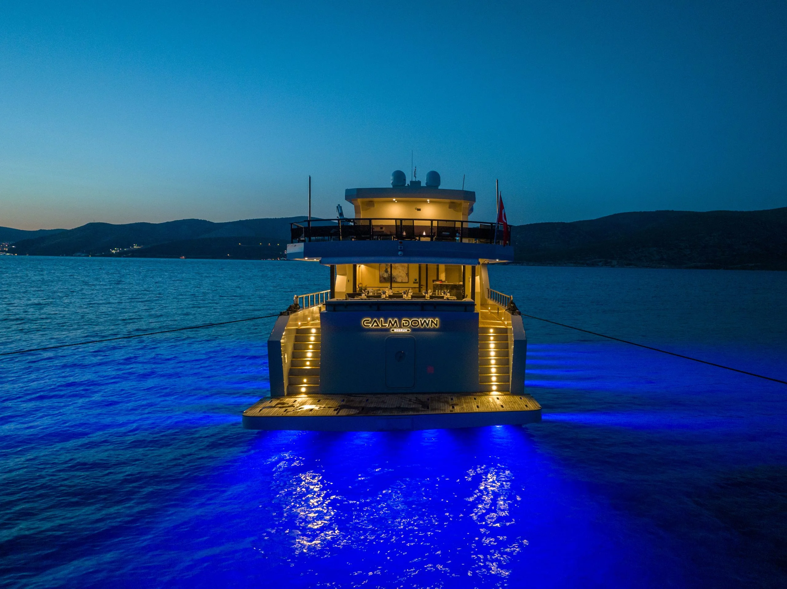 A luxury yacht anchored at dusk, illuminated with bright blue underwater lights, creating a glowing reflection in the calm sea. Hills and a darkening sky are visible in the background.