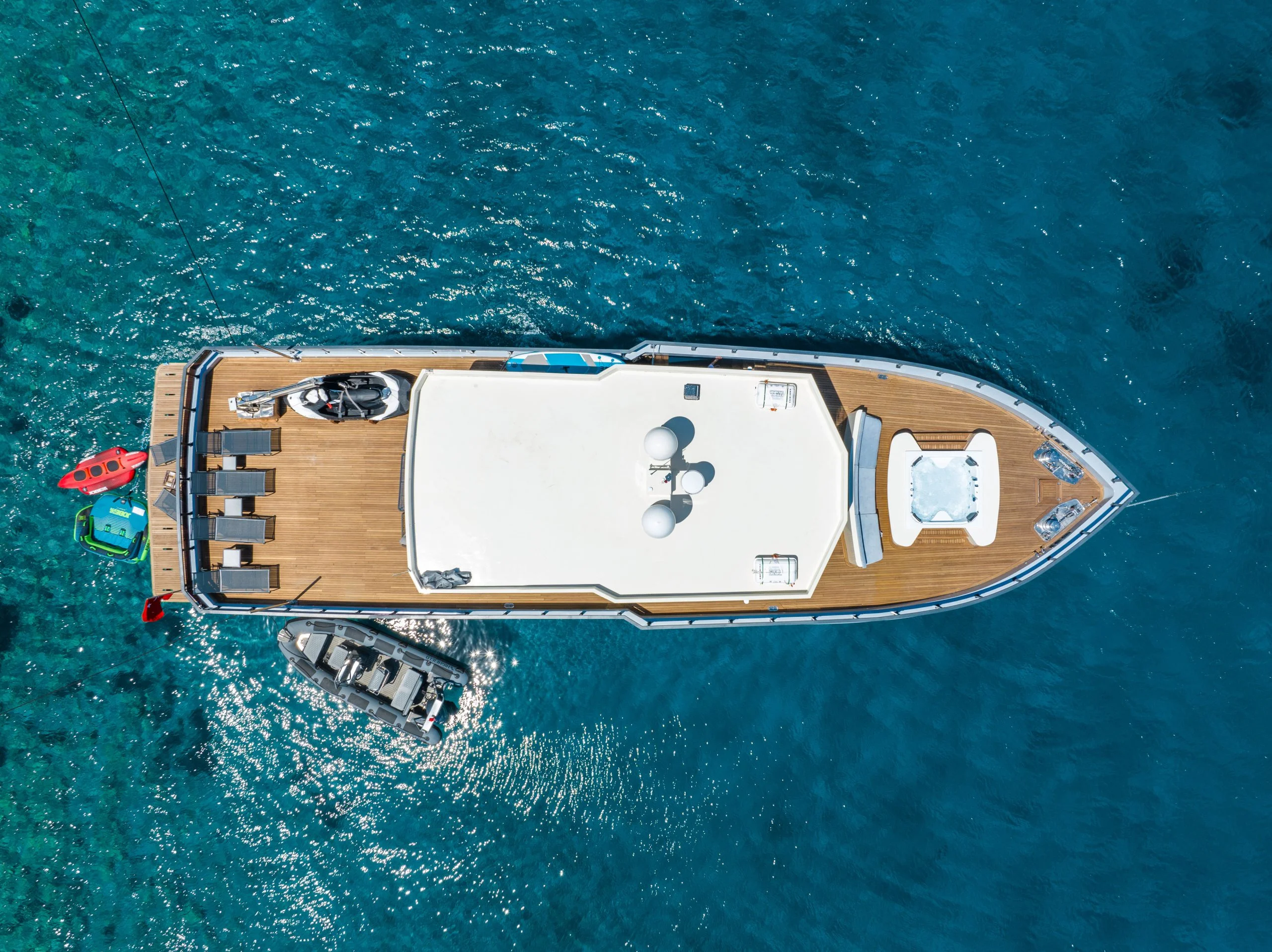 Aerial view of a large yacht on clear blue water, with sun loungers, a hot tub, water scooters, and small boats attached on one side. Sunlight sparkles on the sea around the yacht.