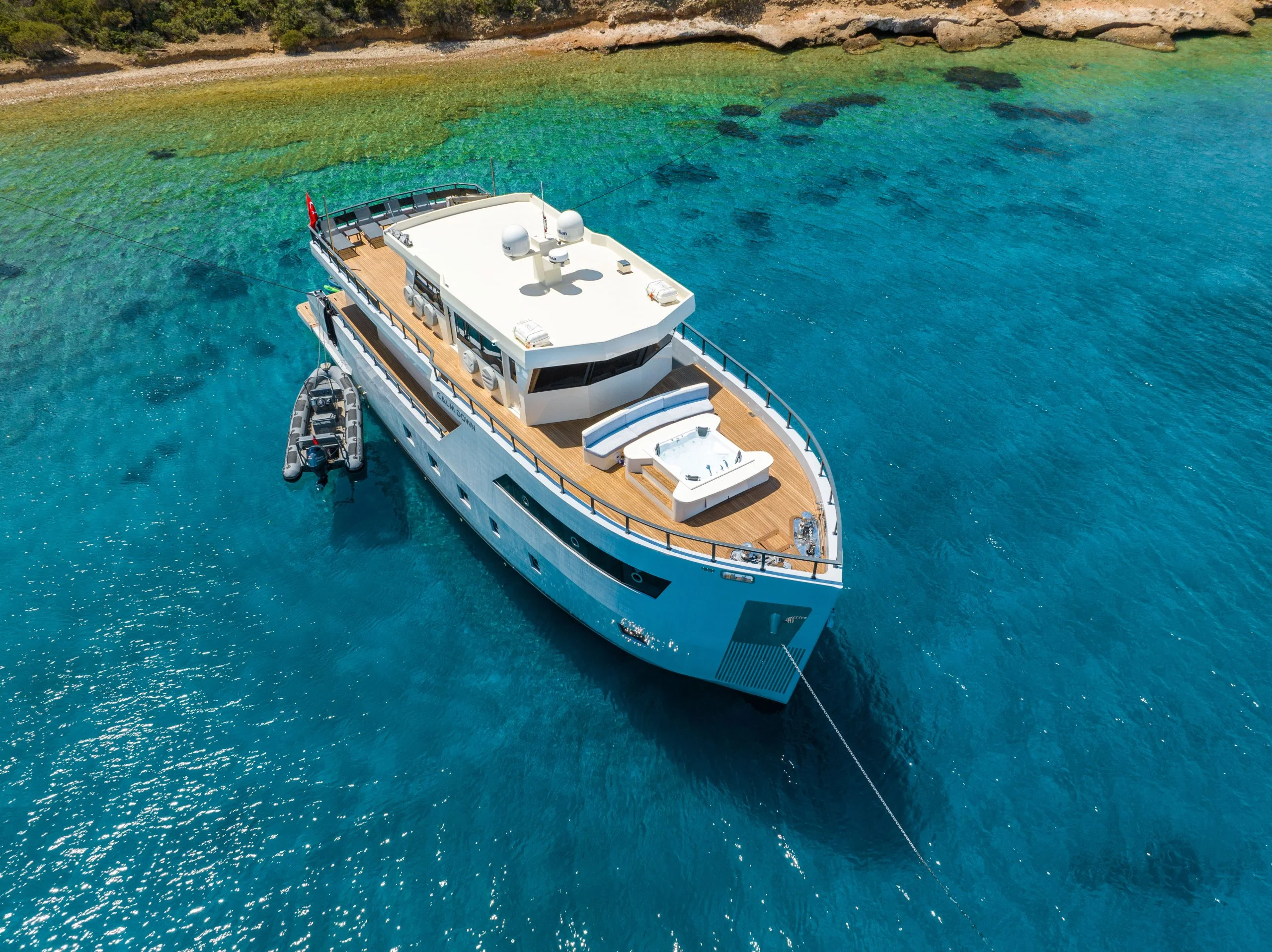 A large white yacht anchored in clear blue water near a rocky shoreline, with a small inflatable boat tied alongside and sun loungers on the yacht’s deck.