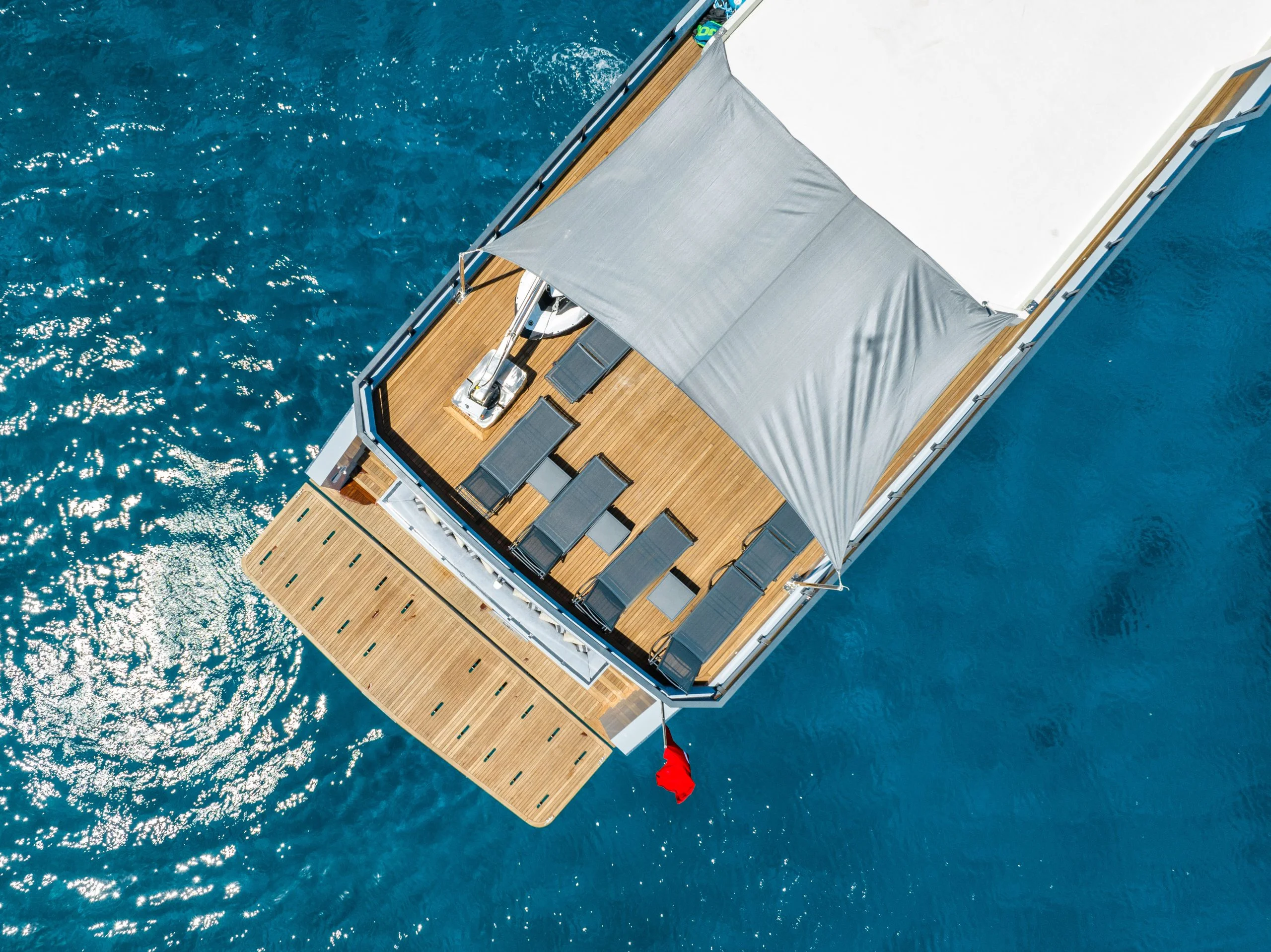 Aerial view of a yacht’s deck with several lounge chairs under a canopy, floating on clear blue water. A small red flag hangs off the back, and sunlight sparkles on the water’s surface.