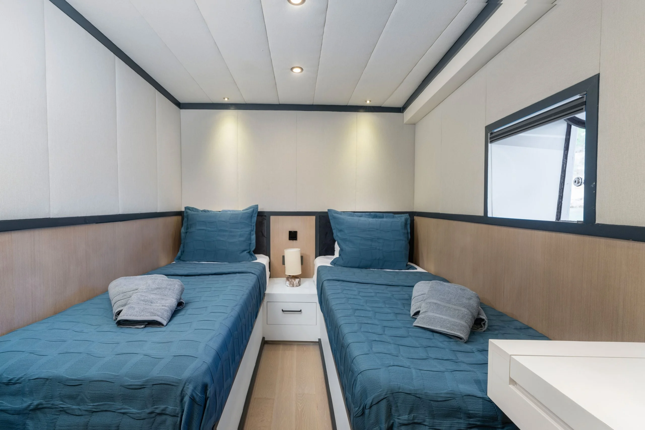 Minimalist bedroom with two single beds, each with teal bedding and pillows. Folded towels are placed on each bed, and there is a small nightstand with a lamp between the beds. A window is on the right wall.
