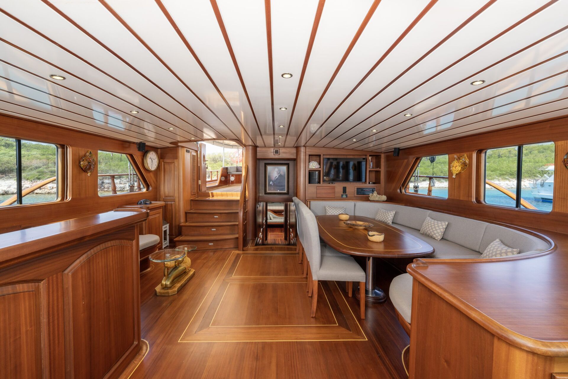 Luxurious yacht interior with polished wood paneling, curved gray seating around a dining table, bar area, and large windows providing views of water and greenery outside.