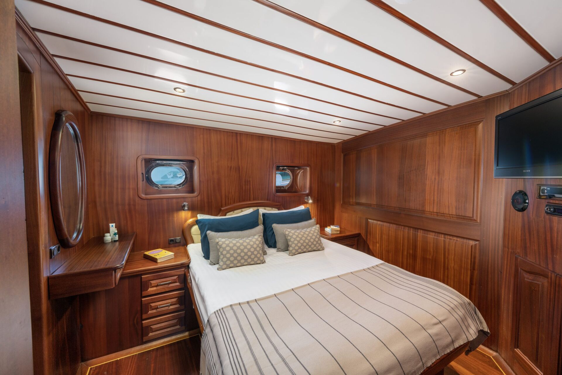 A cozy yacht cabin with wooden paneled walls and ceiling, a double bed with white linens and decorative pillows, small windows, a wall-mounted TV, and a vanity with a round mirror.