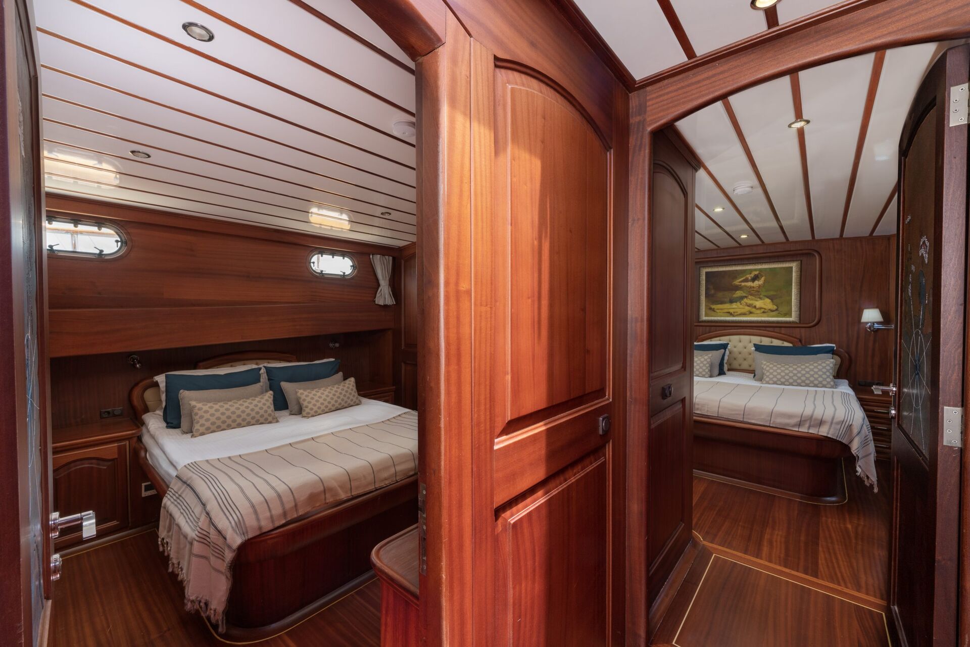 Two adjacent bedrooms on a boat or yacht, each with a neatly made double bed, striped bedding, wooden walls and ceilings, and soft lighting. A painting hangs above one bed; porthole windows provide natural light.