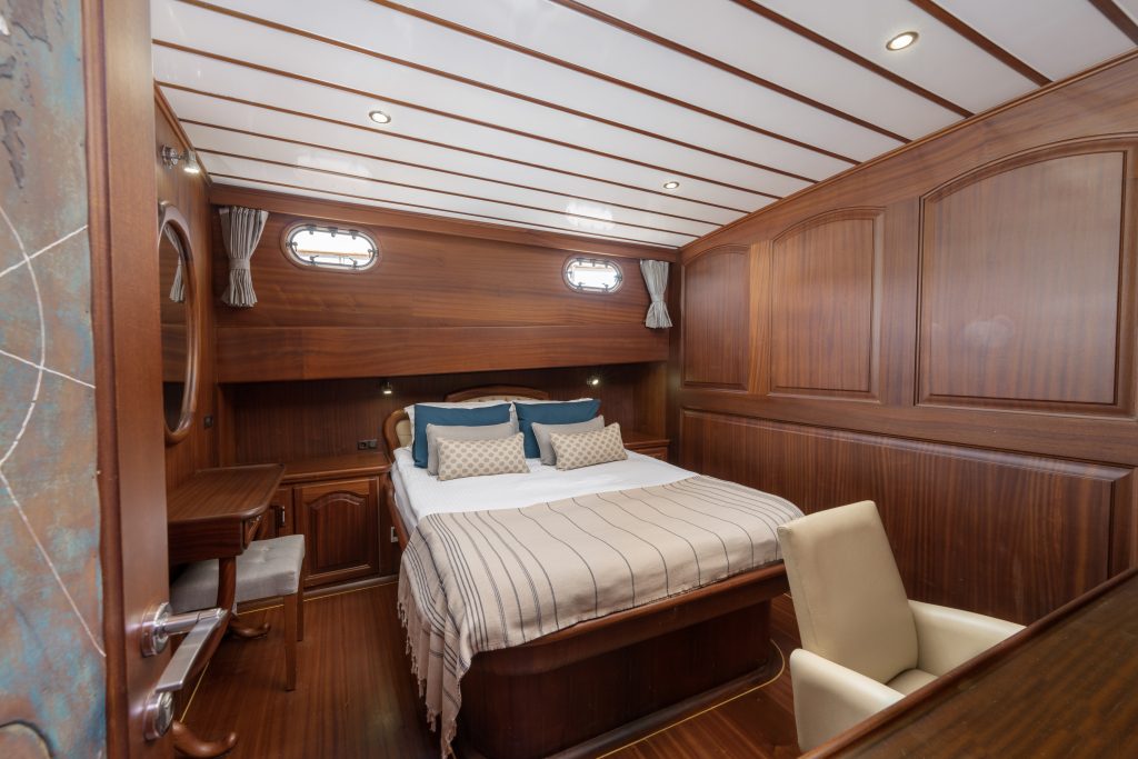 A cozy yacht cabin with wood-paneled walls, a neatly made double bed with striped bedding, decorative pillows, small windows, a desk with a mirror, and a beige chair. Warm lighting creates an inviting atmosphere.