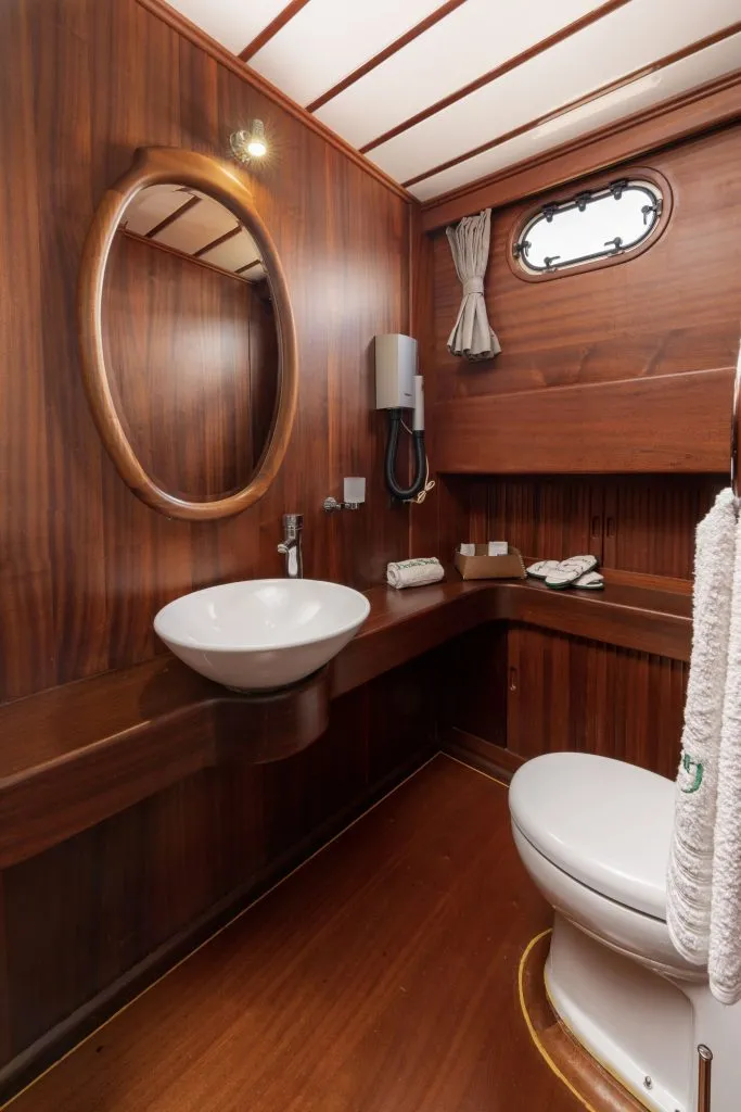 A small, elegant bathroom with wood-paneled walls and floor, a round mirror, a round white sink on a wooden counter, a toilet, a porthole window with curtains, and towels neatly arranged.