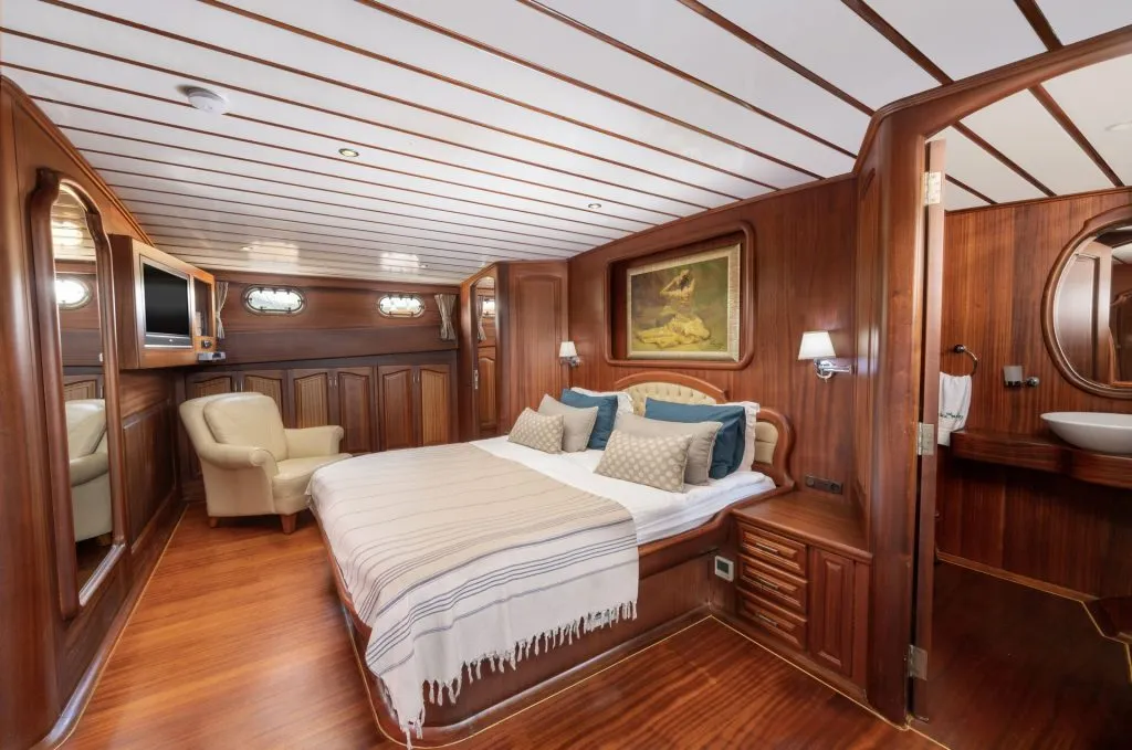 Luxurious yacht cabin with wood-paneled walls and ceiling, a double bed with pillows and a striped blanket, a beige armchair, built-in storage, a wall painting, TV, and an ensuite bathroom with a round mirror.