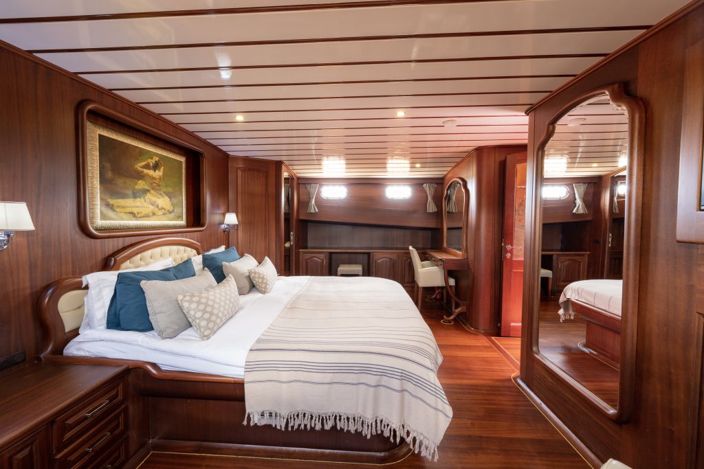 Spacious yacht bedroom with wood-paneled walls, a large bed with white and blue bedding, throw pillows, a painting above the headboard, a desk with a chair, and a mirrored wardrobe reflecting the room.