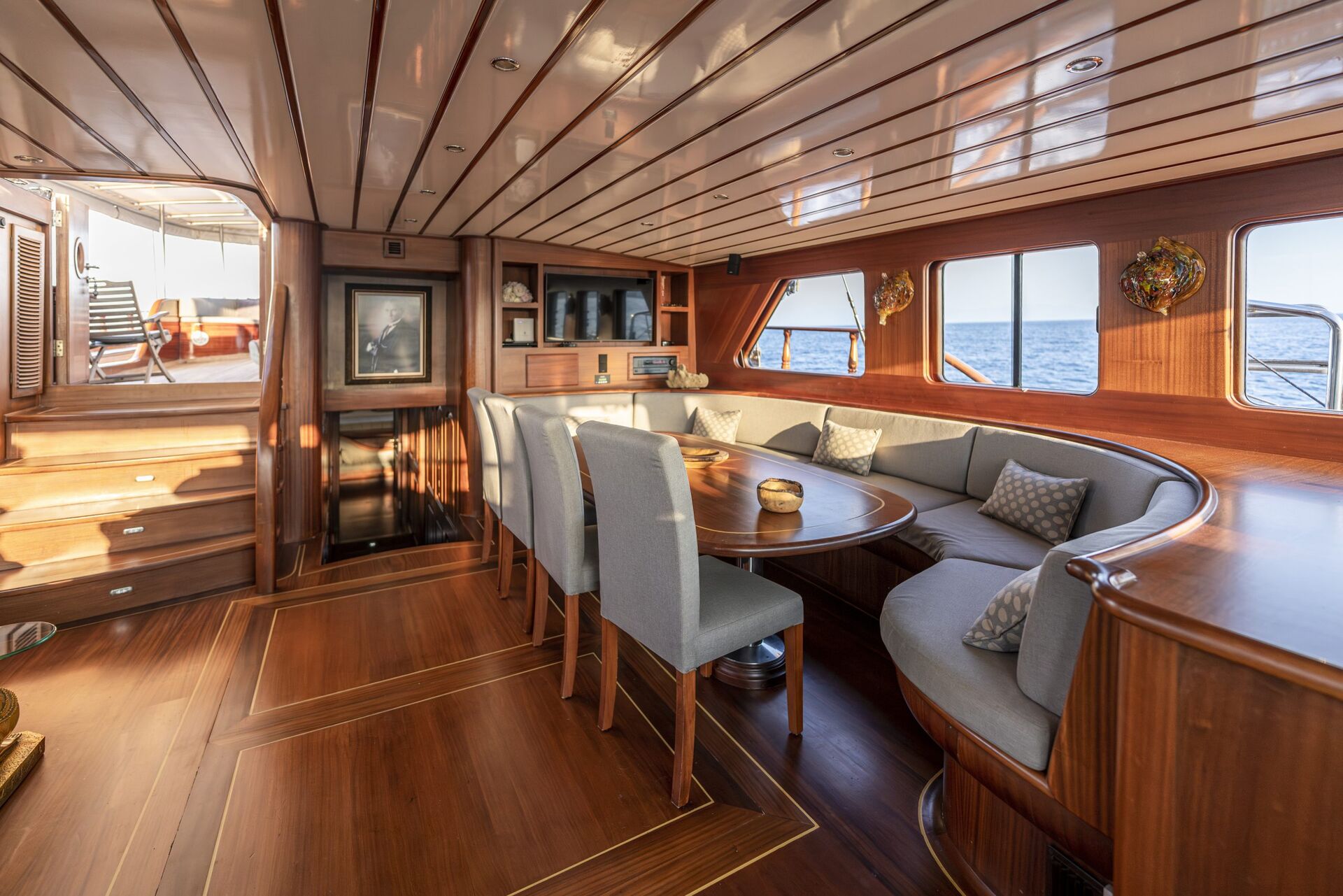 A luxurious yacht interior with polished wooden floors and ceiling, a round dining table surrounded by upholstered chairs and a curved sofa, built-in shelves, large windows, and an ocean view.