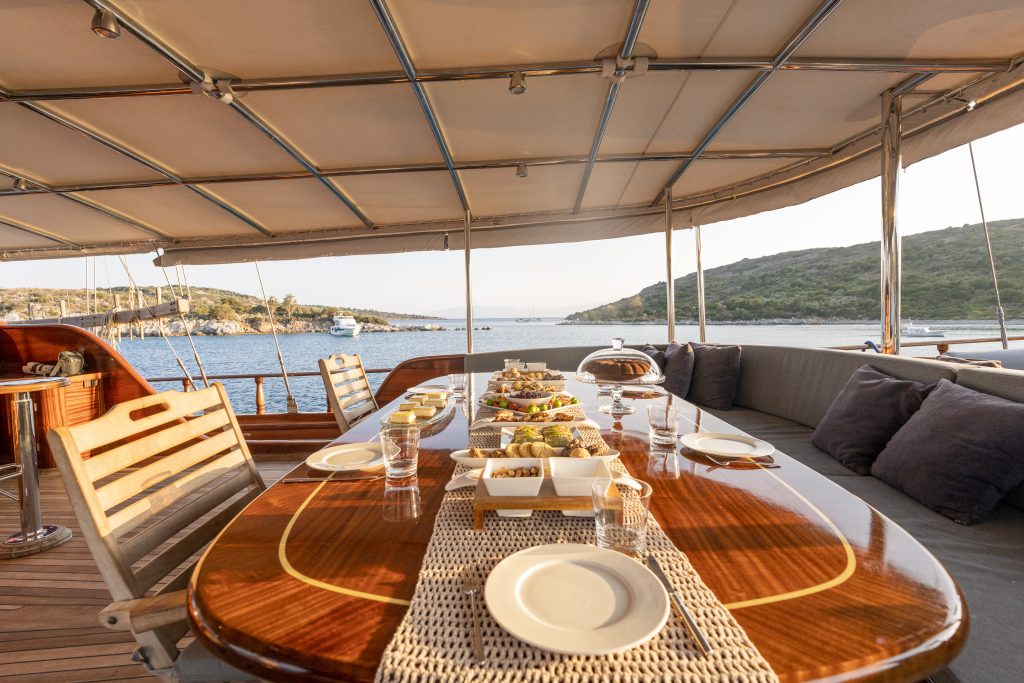 A wooden dining table on a yacht is set with plates, glasses, and various dishes, overlooking calm blue water and green hills under a canopy on a sunny day.