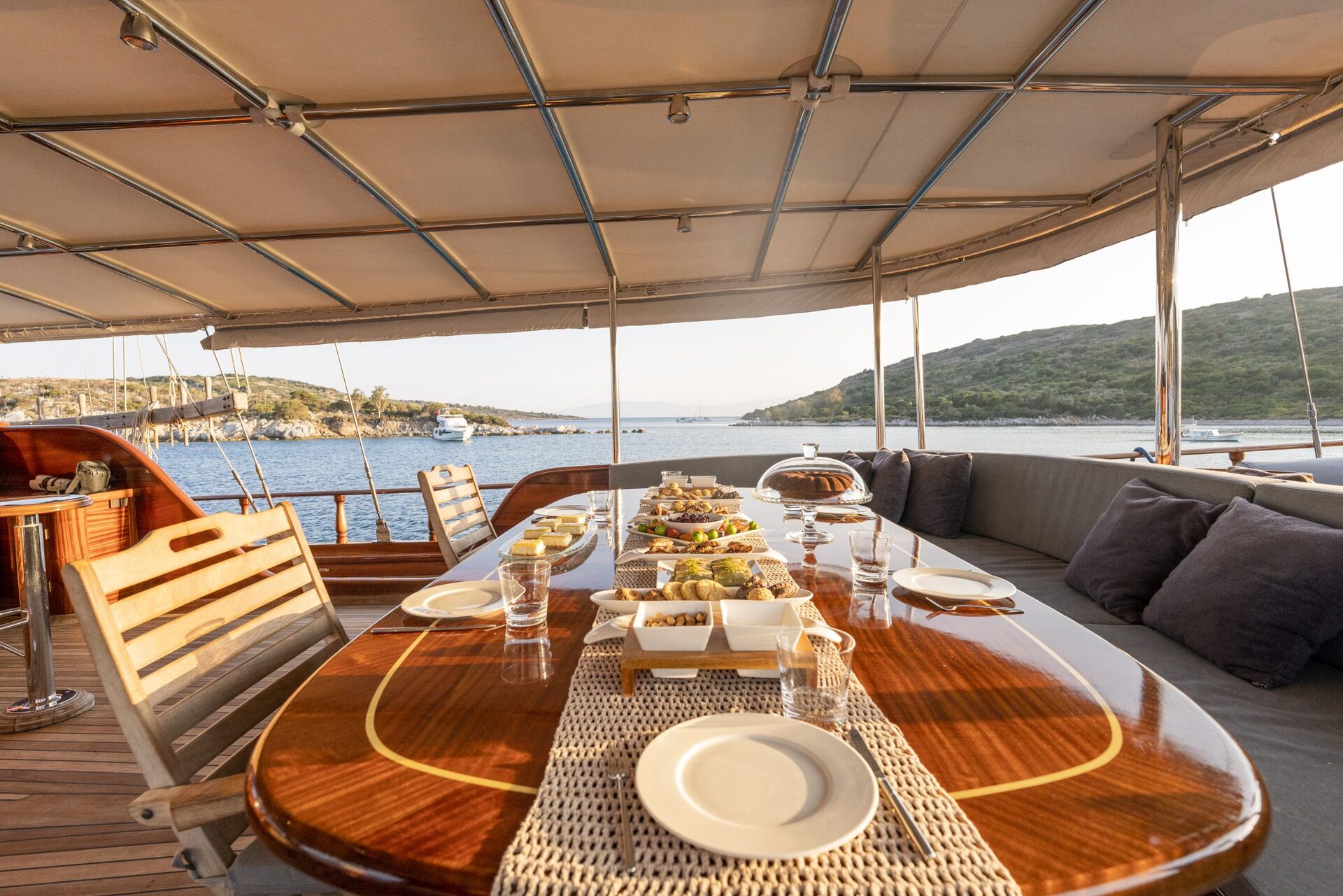 A wooden dining table on a yacht is set with plates, glasses, and various dishes, overlooking calm blue water and green hills under a canopy on a sunny day.