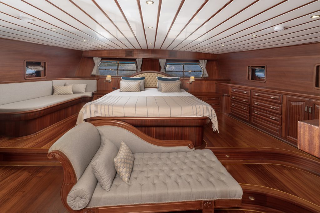 Spacious yacht bedroom with a large bed, wood-paneled walls and floor, built-in drawers, wide windows, a tufted chaise lounge, and a cushioned bench creating a luxurious and elegant atmosphere.