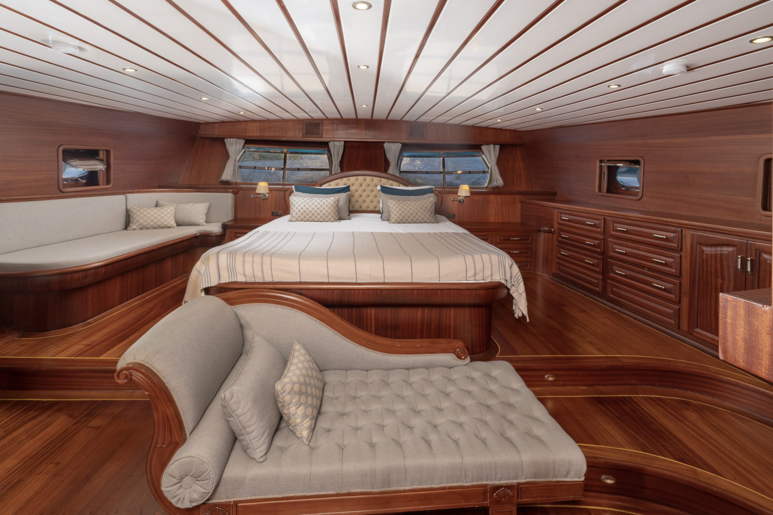 Spacious yacht bedroom with a large bed, wood-paneled walls and floor, built-in drawers, wide windows, a tufted chaise lounge, and a cushioned bench creating a luxurious and elegant atmosphere.