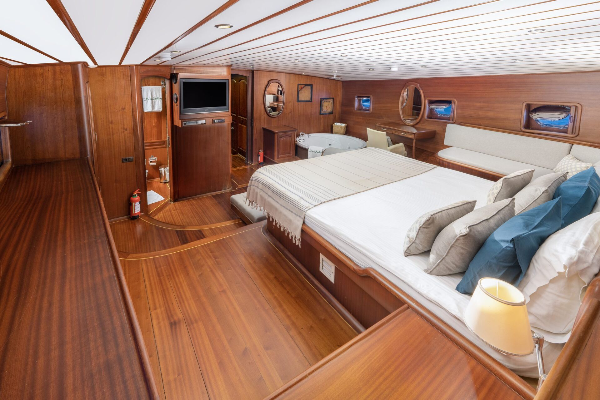 Spacious yacht cabin with a large bed, wooden floors and walls, sofa, flat-screen TV, round windows, and a lamp on a bedside table. Theres also a Jacuzzi tub and various decor accents.