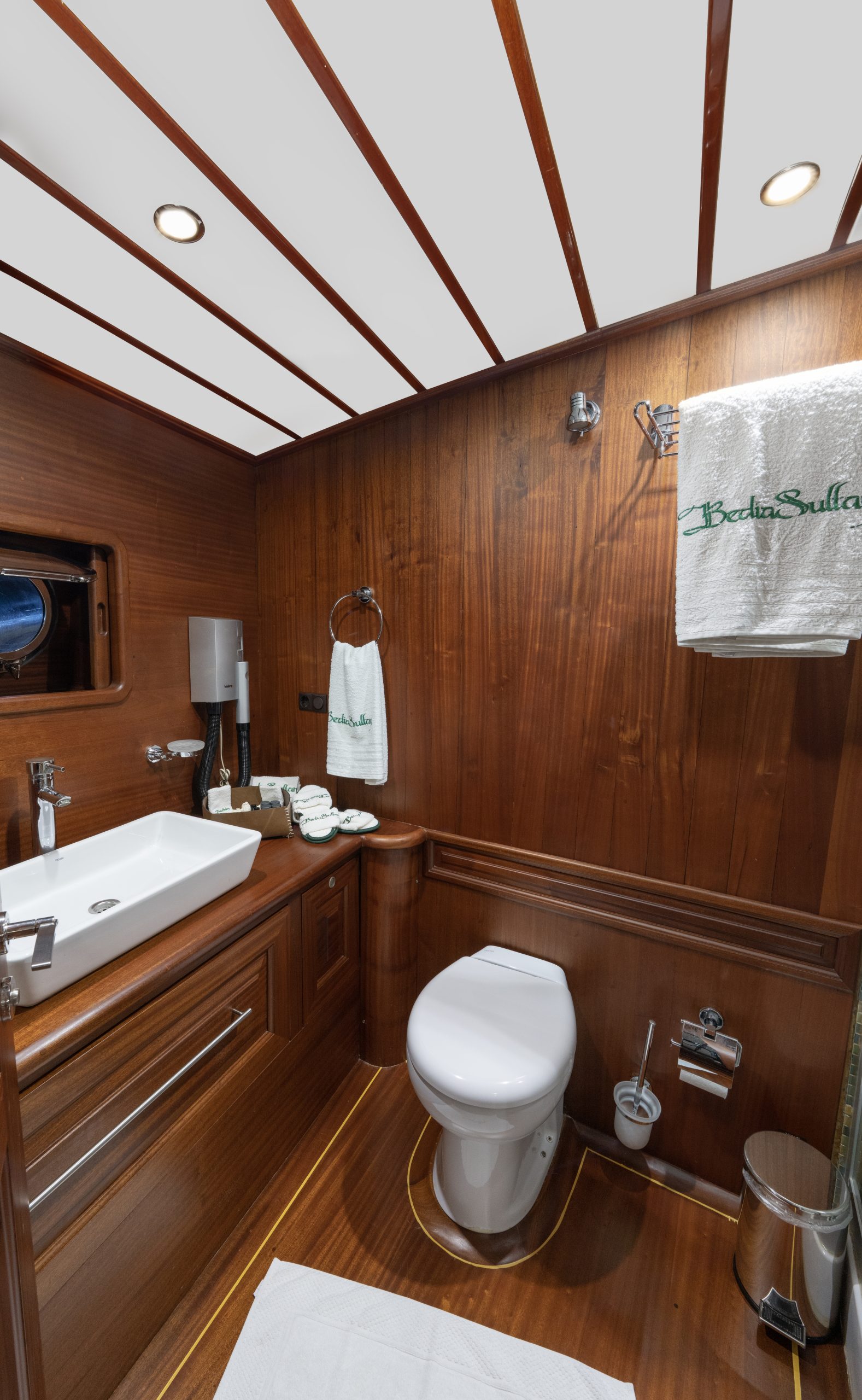 A compact, elegant bathroom with wood-paneled walls and floor, a white toilet, modern sink, towel rack, and amenities. Towels embroidered with Bedia Sultan are neatly hung and folded.