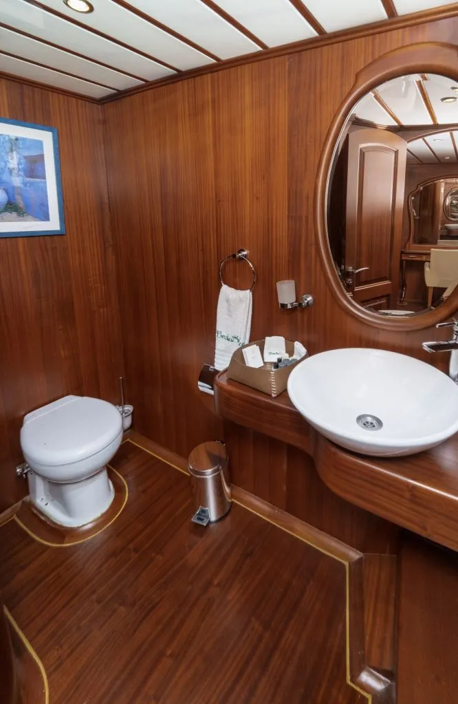 A small bathroom with wooden walls and floor, featuring a white toilet, a round sink on a wooden counter, a round mirror, a towel ring with a towel, a trash can, and a framed picture on the wall.