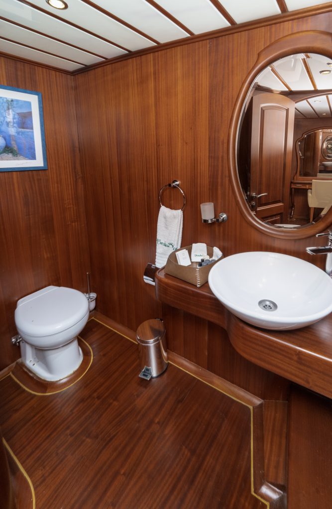 A small bathroom with wooden walls and floor, featuring a white toilet, a round sink on a wooden counter, a round mirror, a towel ring with a towel, a trash can, and a framed picture on the wall.