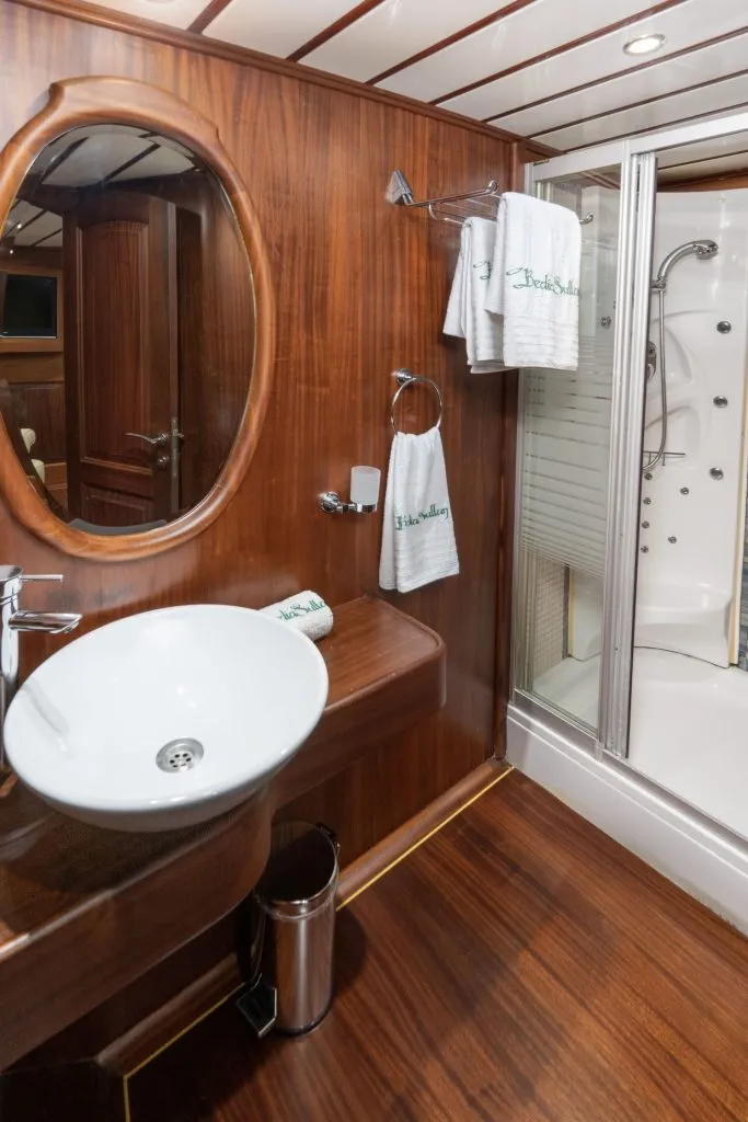 A bathroom with wood-paneled walls and floors, a round mirror above a modern white sink, white towels on racks, a trash bin below the sink, and a glass-enclosed shower in the corner.