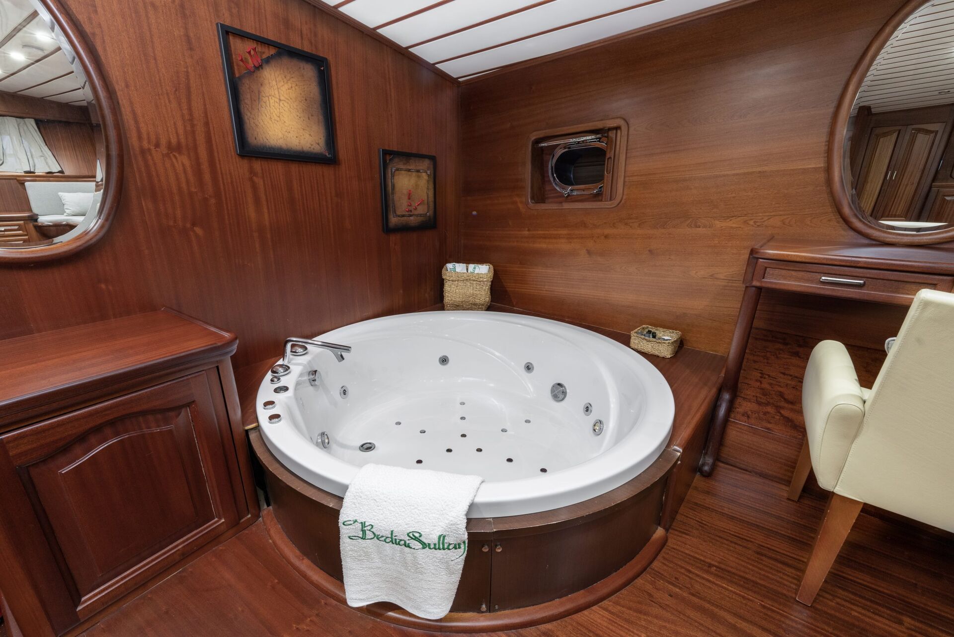 A luxurious round jacuzzi tub with jets, set in a wooden paneled room. A white towel hangs on the tub, and a small desk with a cream chair is nearby. Framed art decorates the walls.