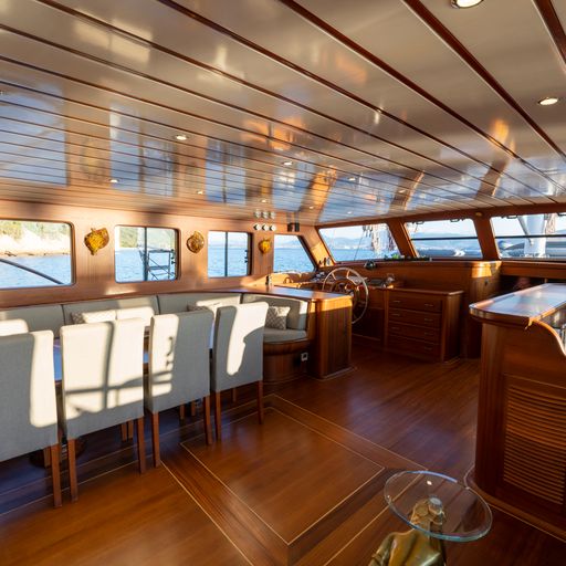 Spacious yacht interior with polished wood floors and ceiling, a dining table with white chairs, large windows letting in sunlight, and a steering area at the front.