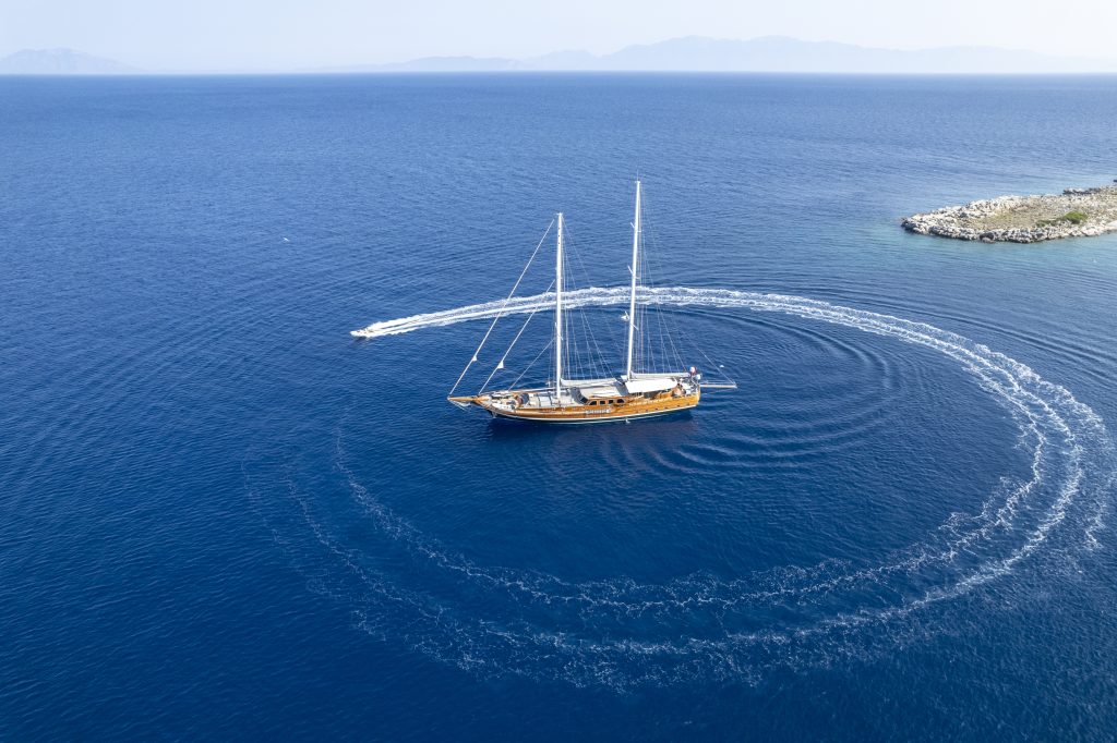 Aerial view of a sailboat anchored in calm blue water with a speedboat circling it, leaving white wake trails in a perfect ring. A small rocky coastline is visible to the right.