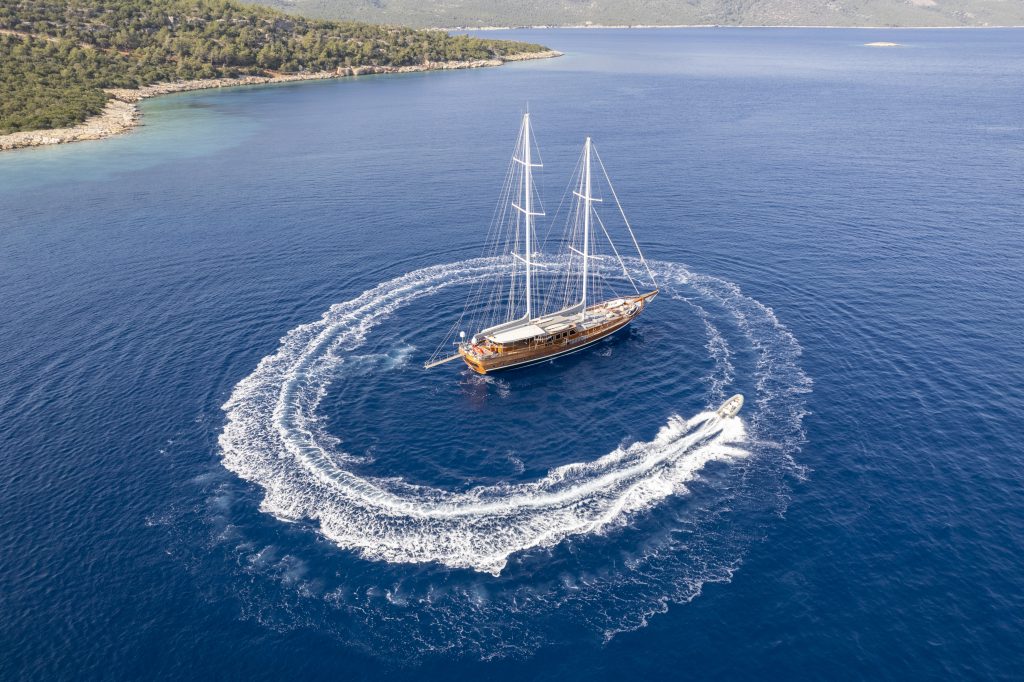 Aerial view of a large sailboat anchored in blue water as a small speedboat circles around it, leaving a white wake, with a forested shoreline in the background.