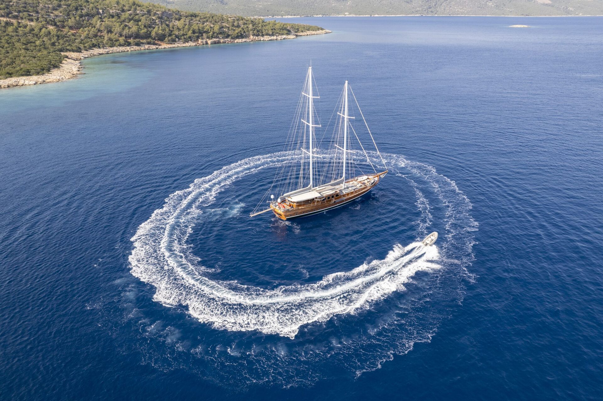 Aerial view of a large sailboat anchored in blue water as a small speedboat circles around it, leaving a white wake, with a forested shoreline in the background.