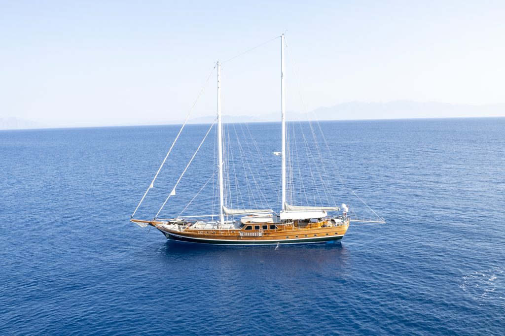A large wooden sailboat with two masts floats on calm, blue ocean water under a clear sky, with distant mountains faintly visible on the horizon.