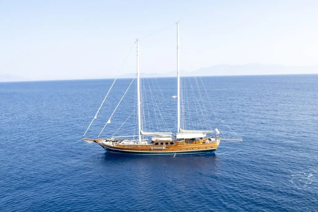 A large wooden sailboat with two masts floats on calm, blue ocean water under a clear sky, with distant mountains faintly visible on the horizon.
